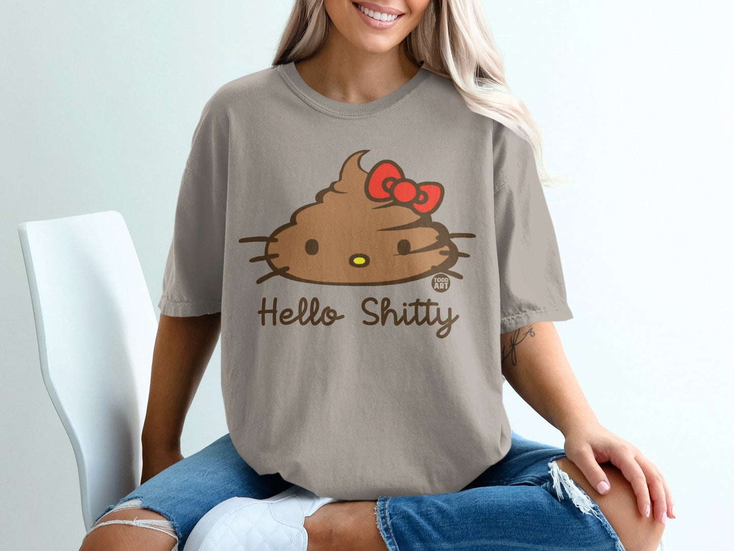 Brown poop emoji with red bow and "Hello Shitty" text on a tan t-shirt.