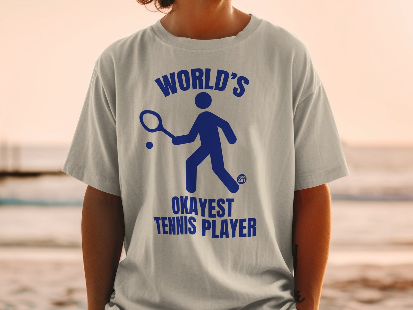 Blue t-shirt with graphic of a person playing tennis and text "WORLD'S OKAYEST TENNIS PLAYER".