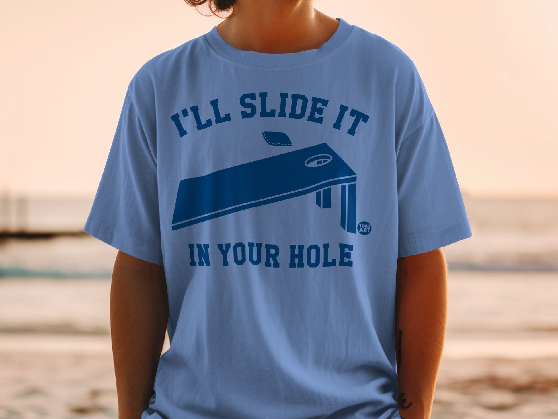 Blue t-shirt with cornhole board graphic and text.