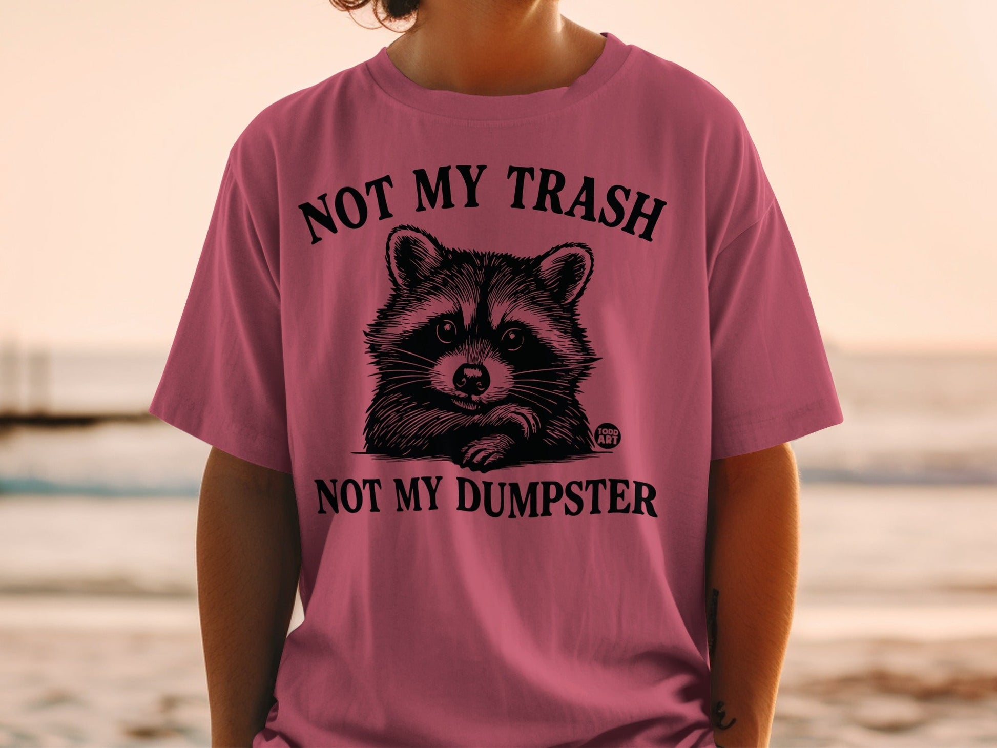 Pink t-shirt with raccoon graphic and text: "Not My Trash, Not My Dumpster".