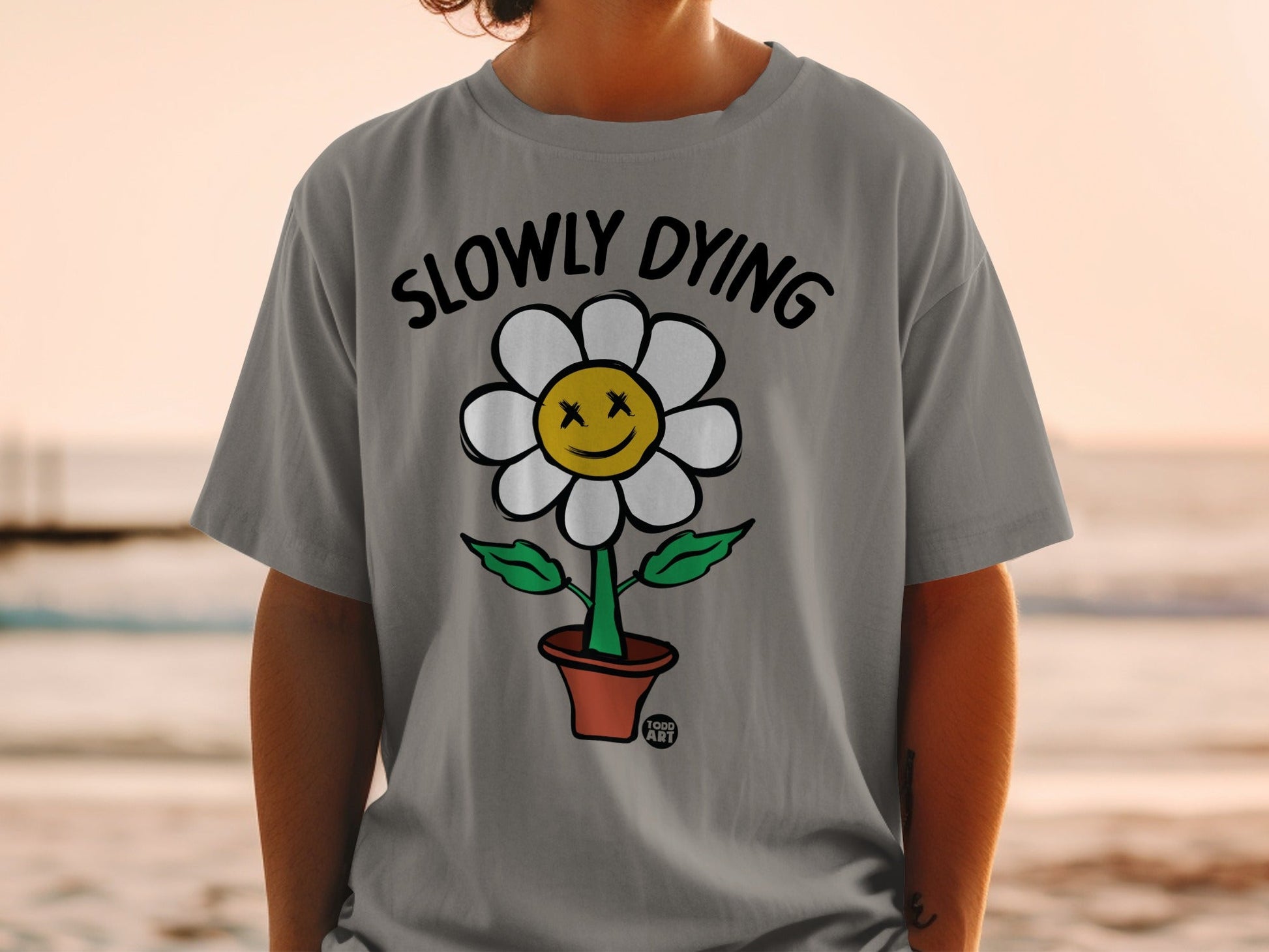 Gray t-shirt with a cartoon flower wearing a smiley face and the text "SLOWLY DYING".
