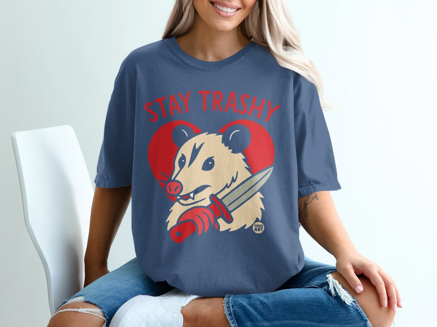 Blue t-shirt with red text "STAY TRASHY" and a possum holding a knife.