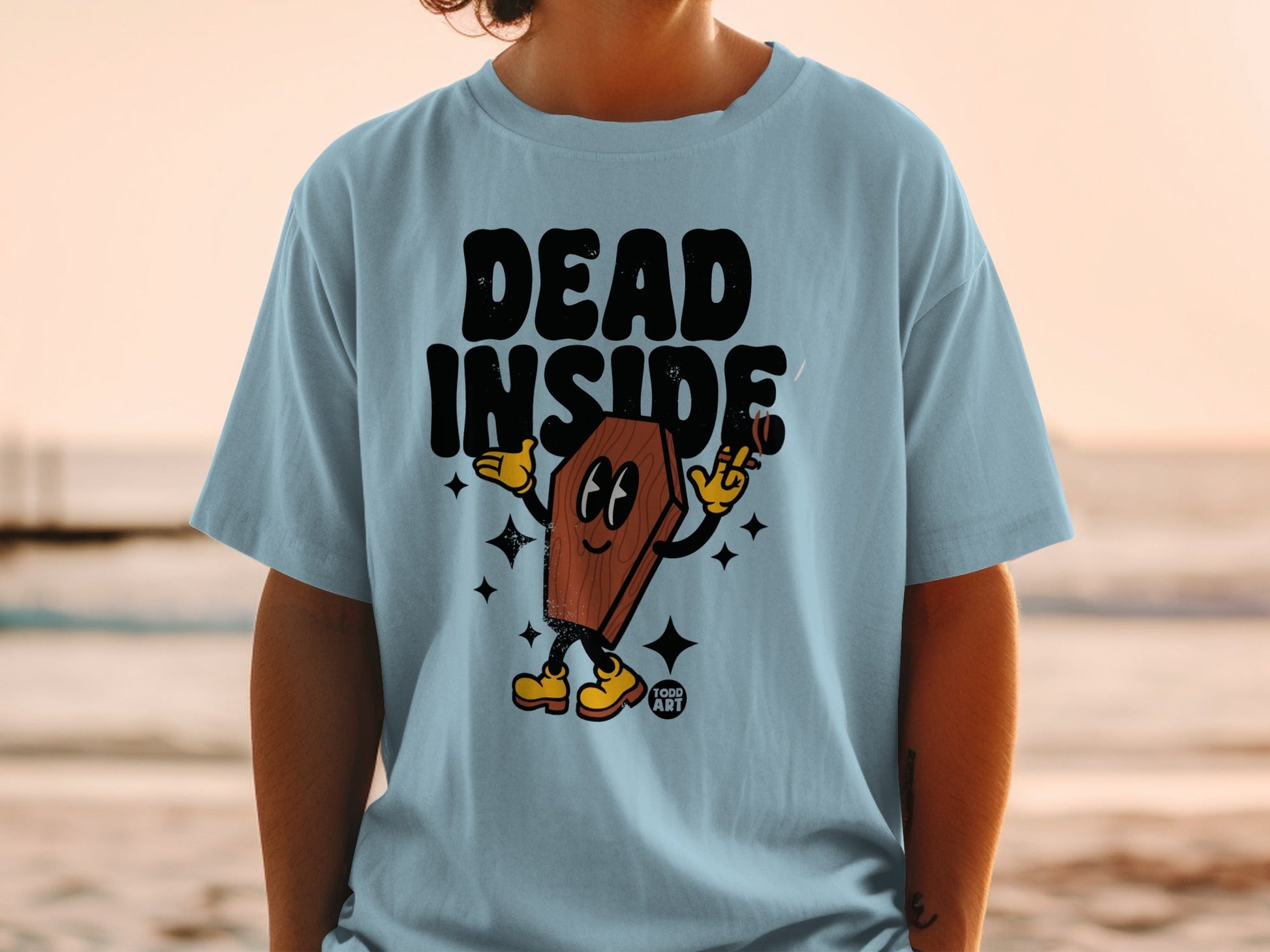 Light blue t-shirt with cartoon coffin character on beach