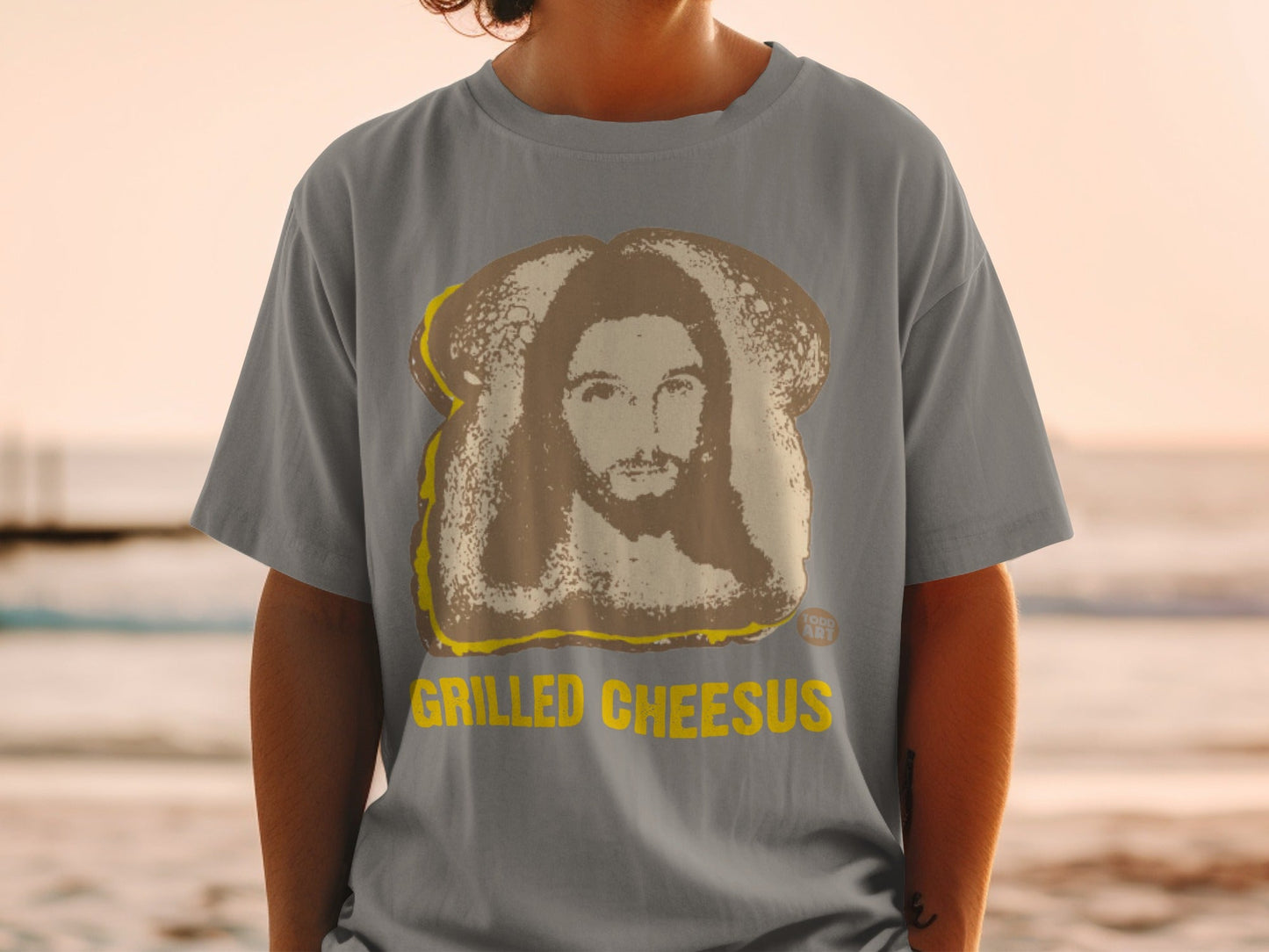 Grilled Cheesus graphic on a gray t-shirt with a beach background.