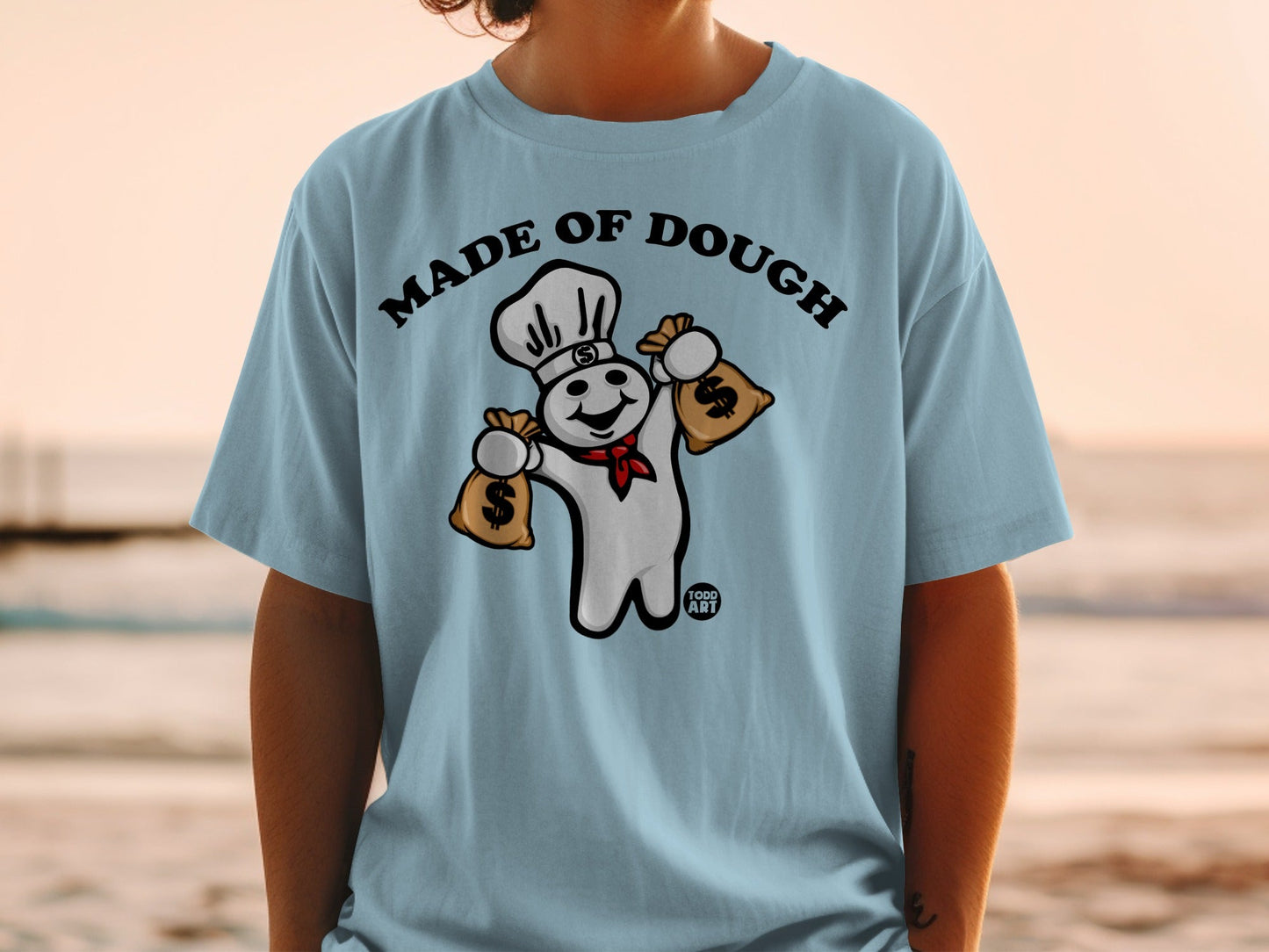 Pillsbury Doughboy wearing chef hat holding money bags on light blue t-shirt.