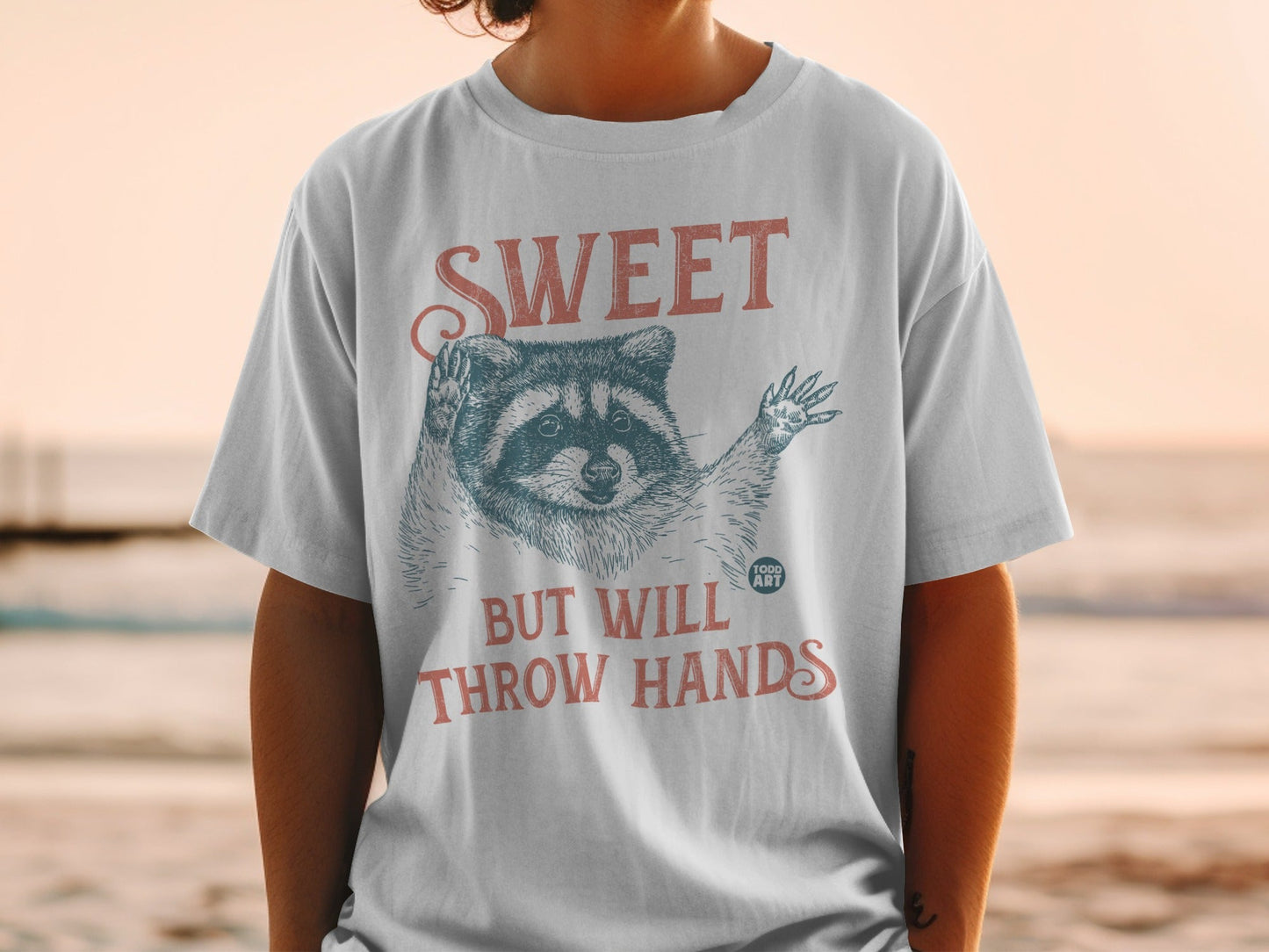T-shirt with raccoon graphic and text "SWEET BUT WILL THROW HANDS".