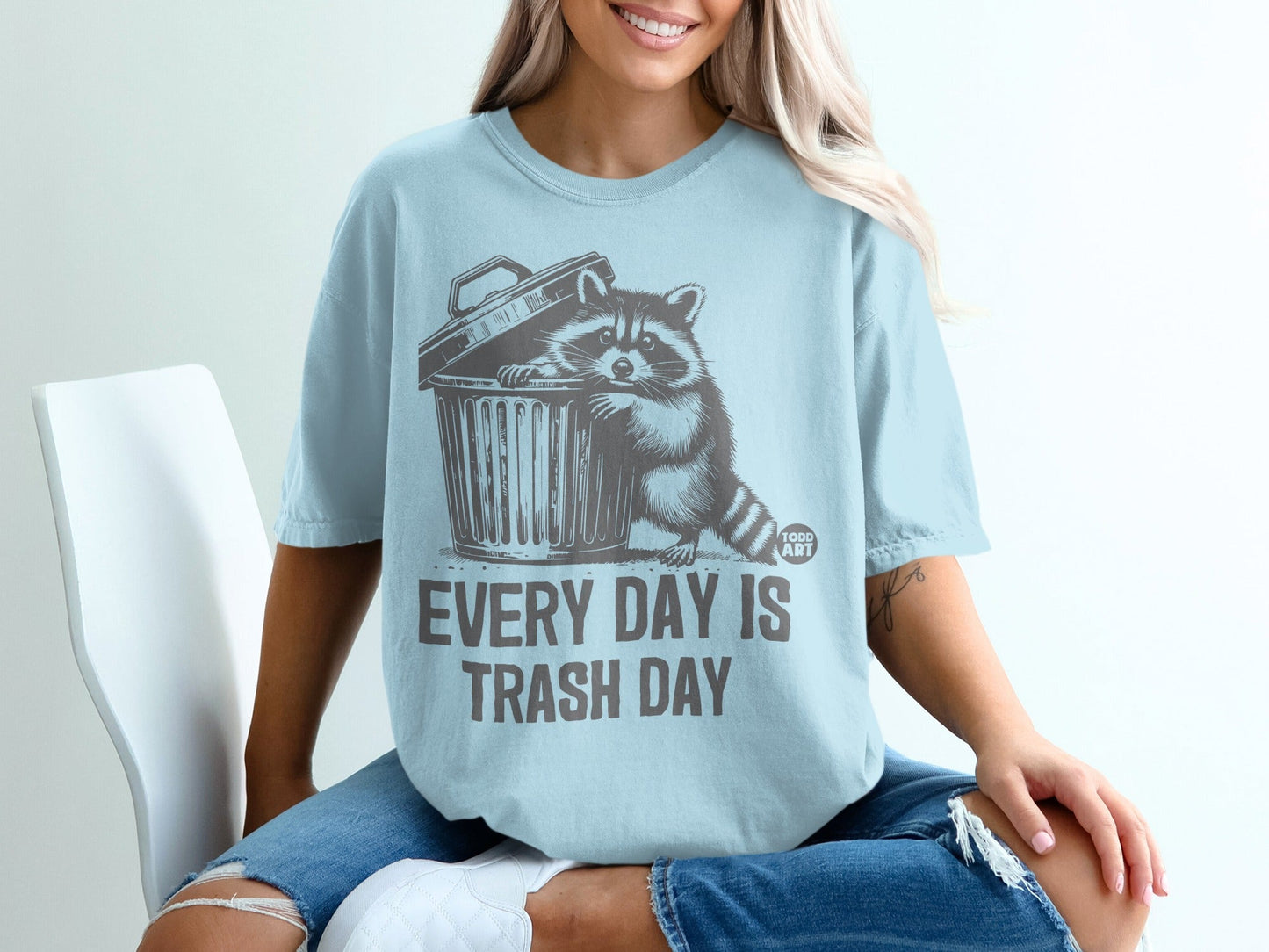 Light blue t-shirt with raccoon peeking out of trash can graphic and text.