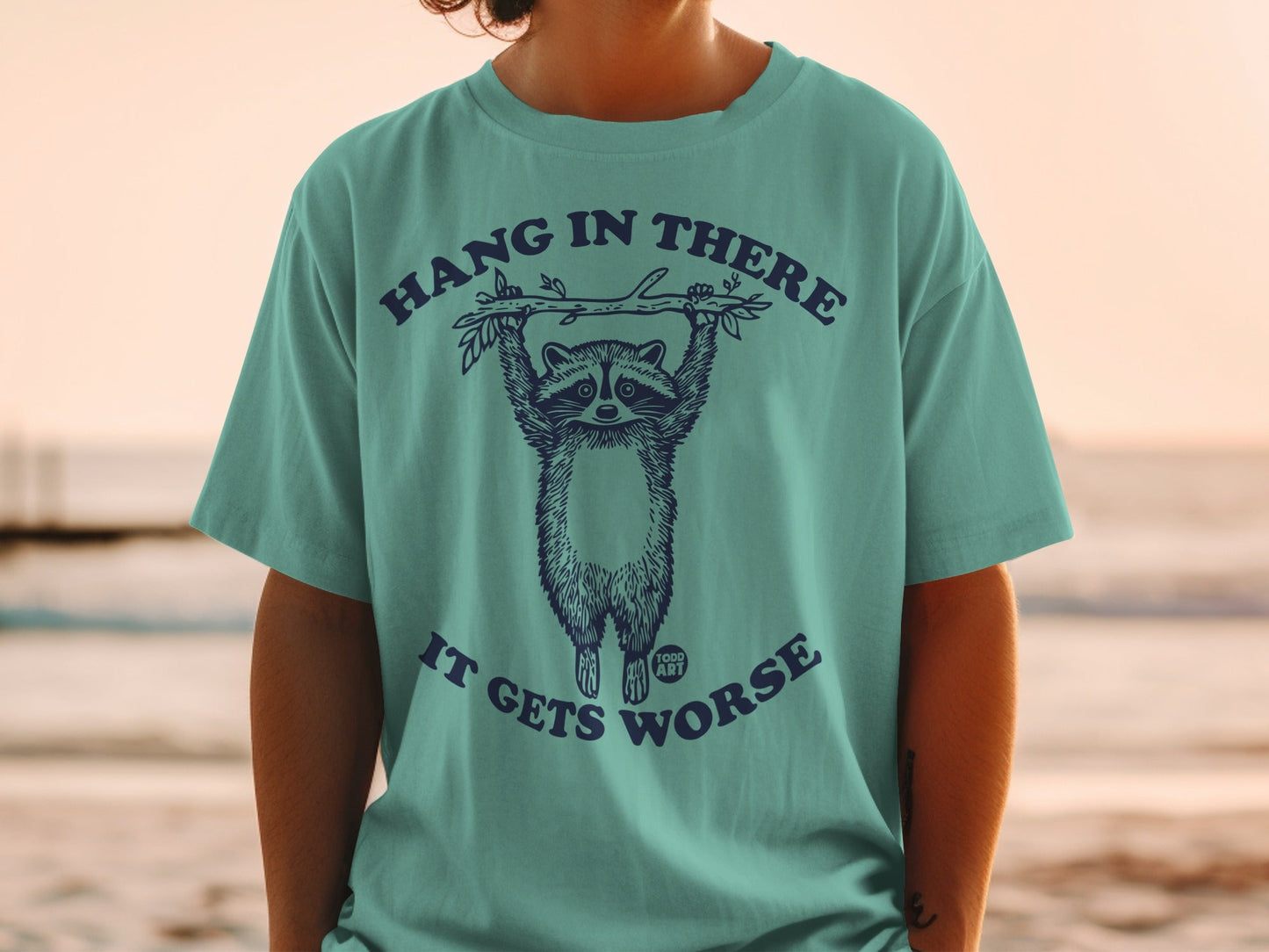 T-shirt with raccoon hanging from a branch saying "Hang in there, it gets worse."