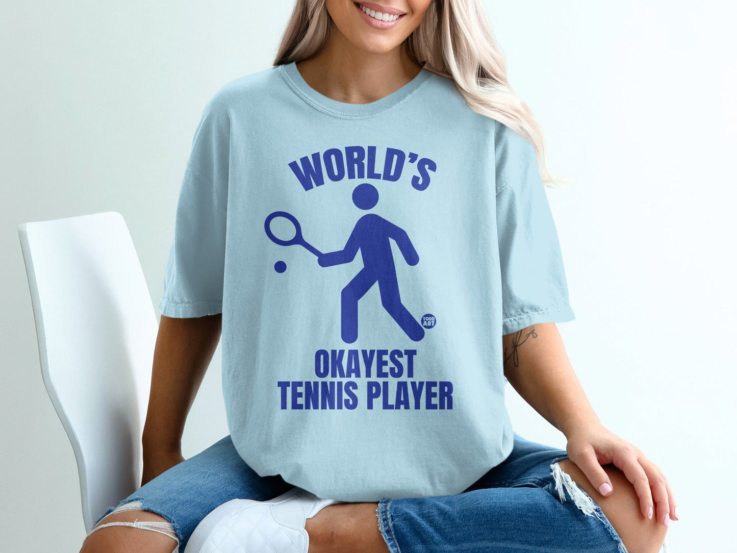 Light blue t-shirt with navy text and graphic: "WORLD'S OKAYEST TENNIS PLAYER".