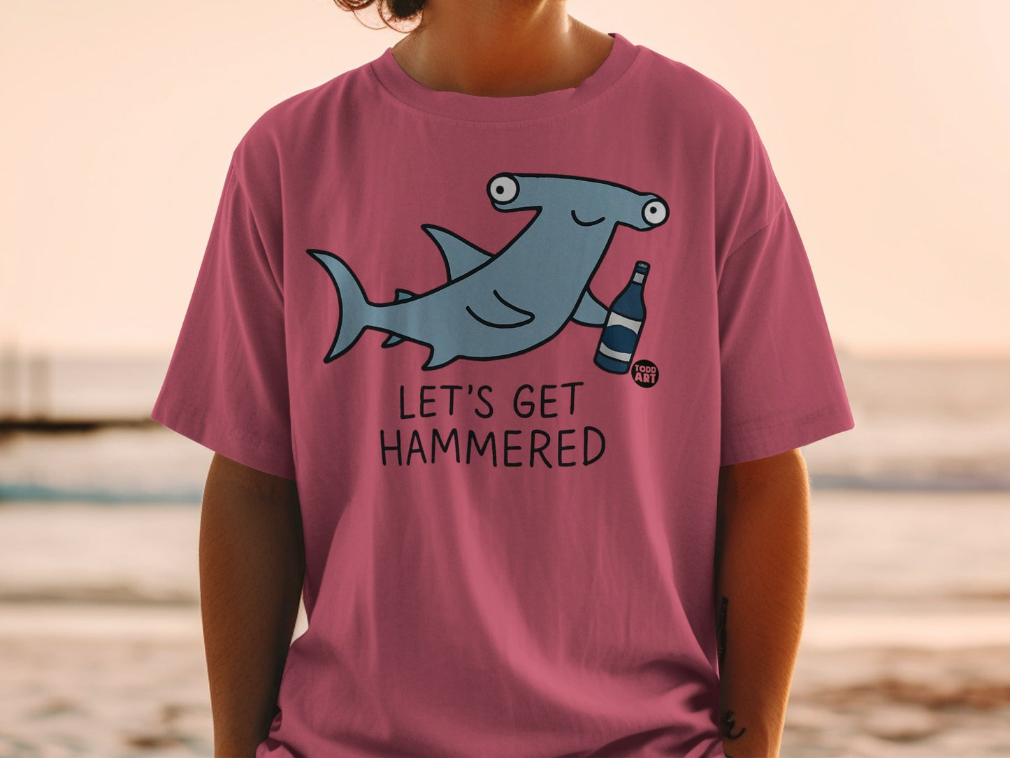 Pink t-shirt with cartoon hammerhead shark holding a bottle, text "LET'S GET HAMMERED".