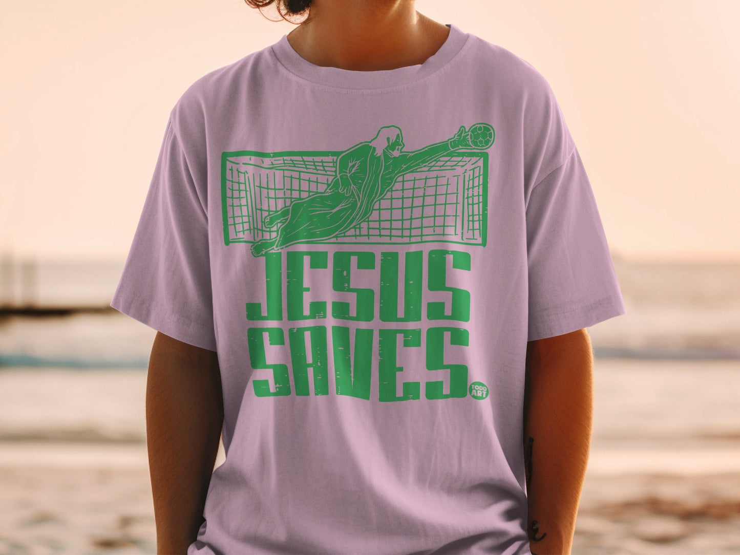 Jesus Saves text with goalkeeper in green on lavender t-shirt.