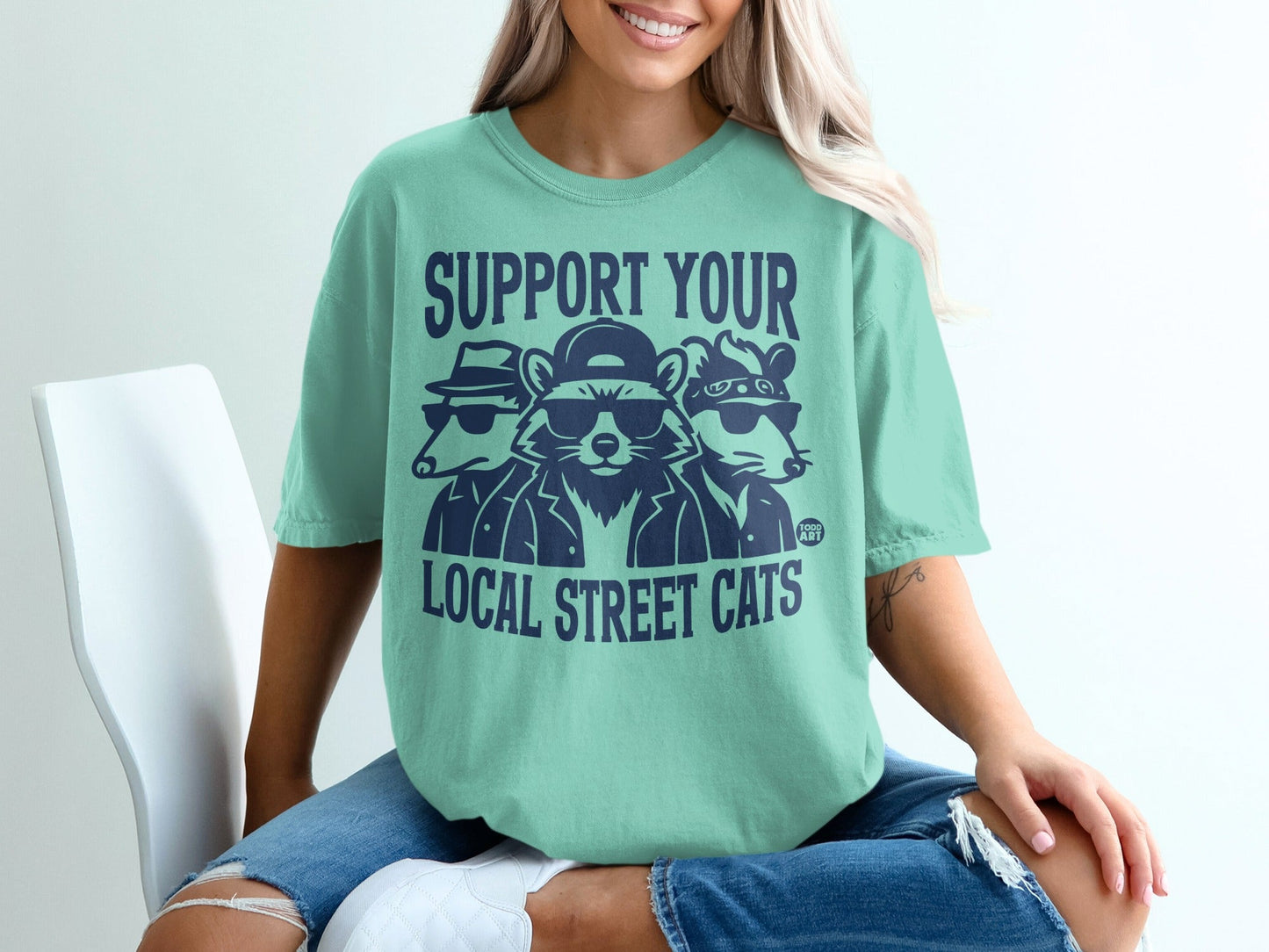 Green t-shirt with cartoon raccoons wearing sunglasses and hats, text "SUPPORT YOUR LOCAL STREET CATS".