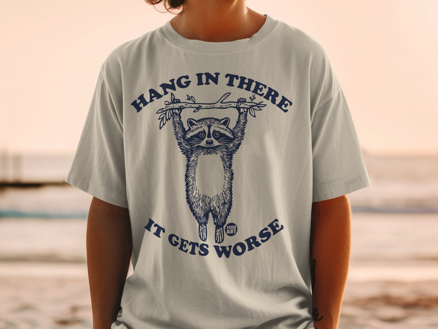 T-shirt with raccoon hanging from branch, text "HANG IN THERE IT GETS WORSE".