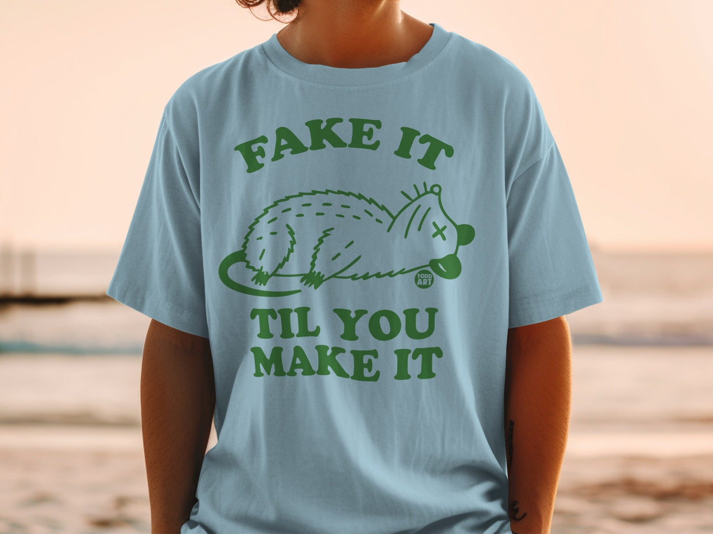 Light blue t-shirt with green text "FAKE IT TIL YOU MAKE IT" and a possum graphic.