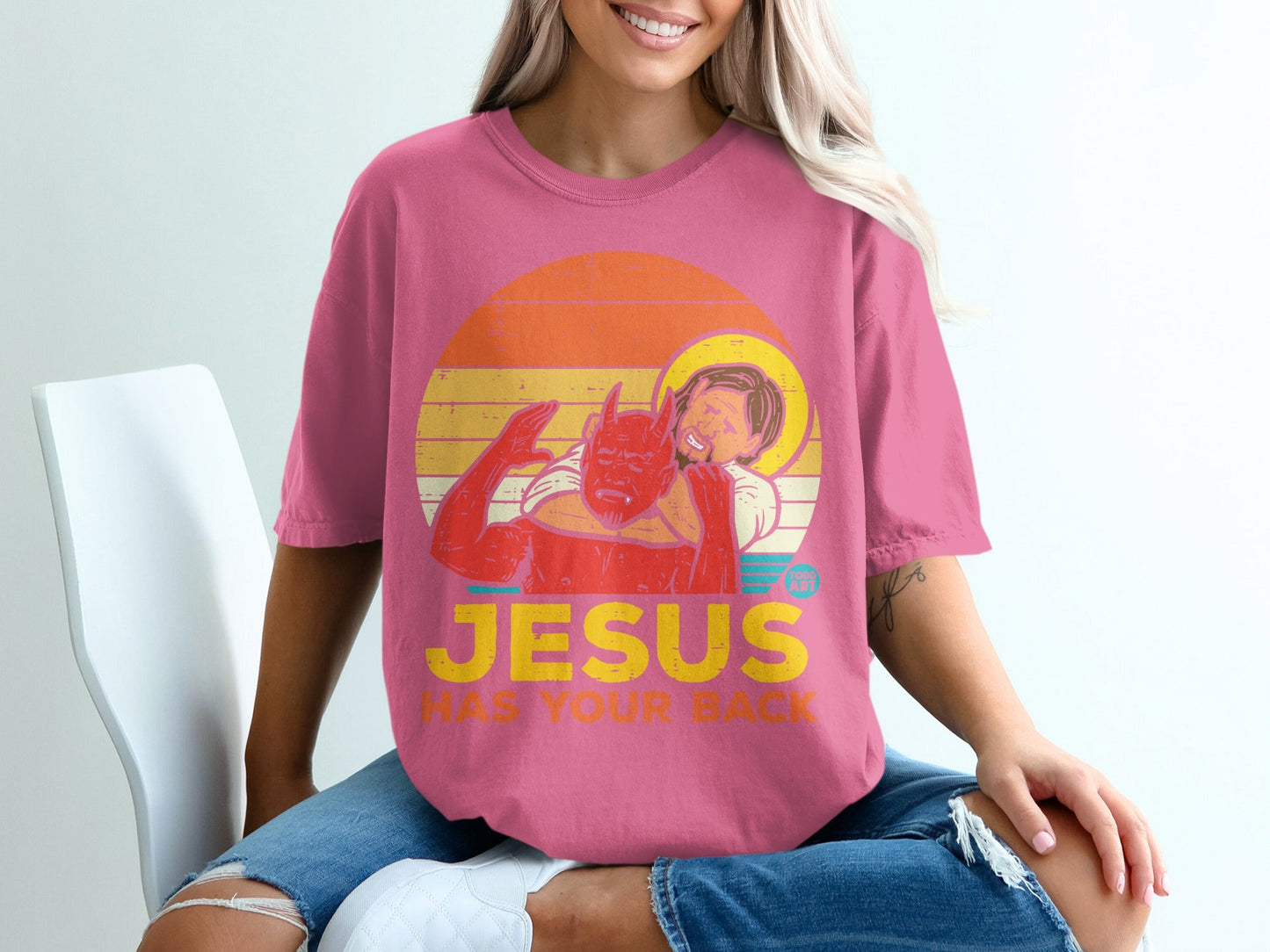 Pink t-shirt with devil hugging Jesus, text "JESUS HAS YOUR BACK."