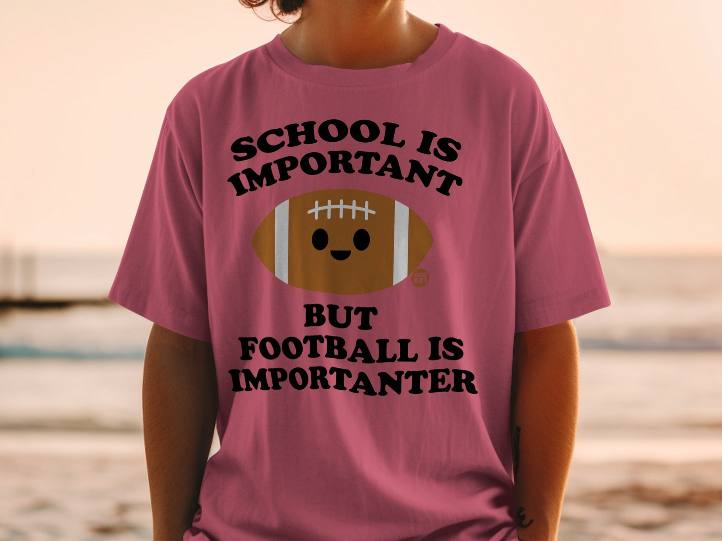 Pink t-shirt with cartoon football graphic and "SCHOOL IS IMPORTANT BUT FOOTBALL IS IMPORTANTER" text.