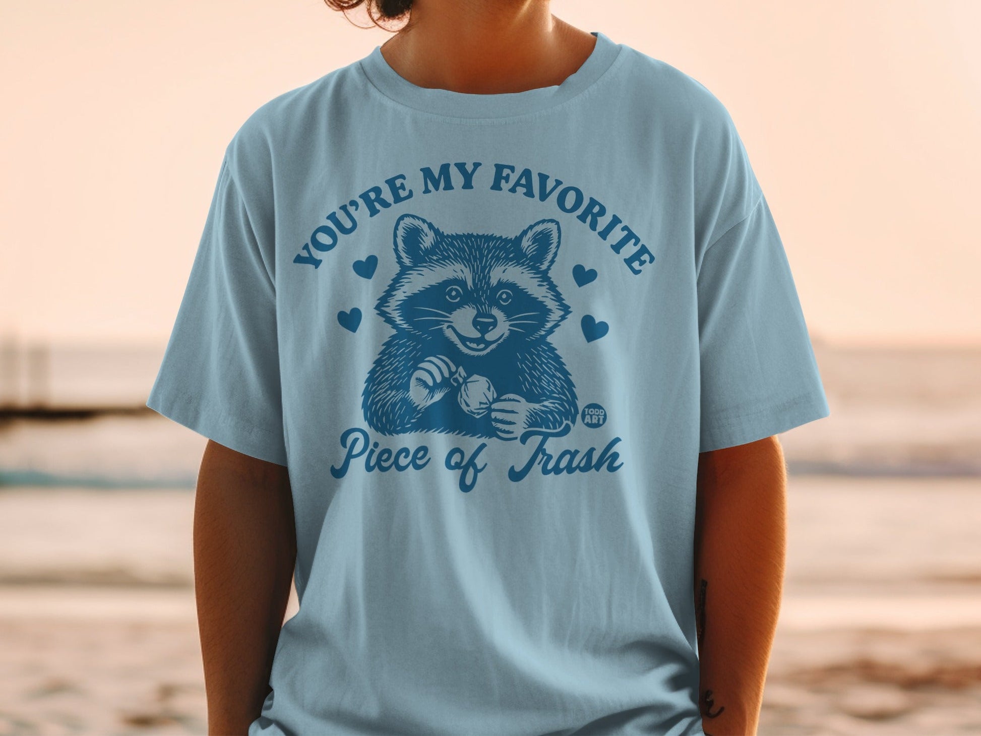 Light blue t-shirt with smiling raccoon and text "YOU'RE MY FAVORITE Piece of Trash".