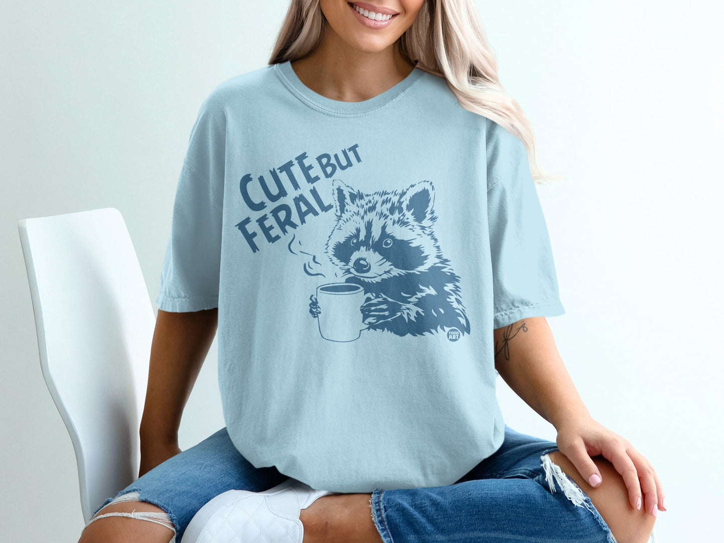 Light blue t-shirt with raccoon drinking coffee graphic, text "Cute but feral".