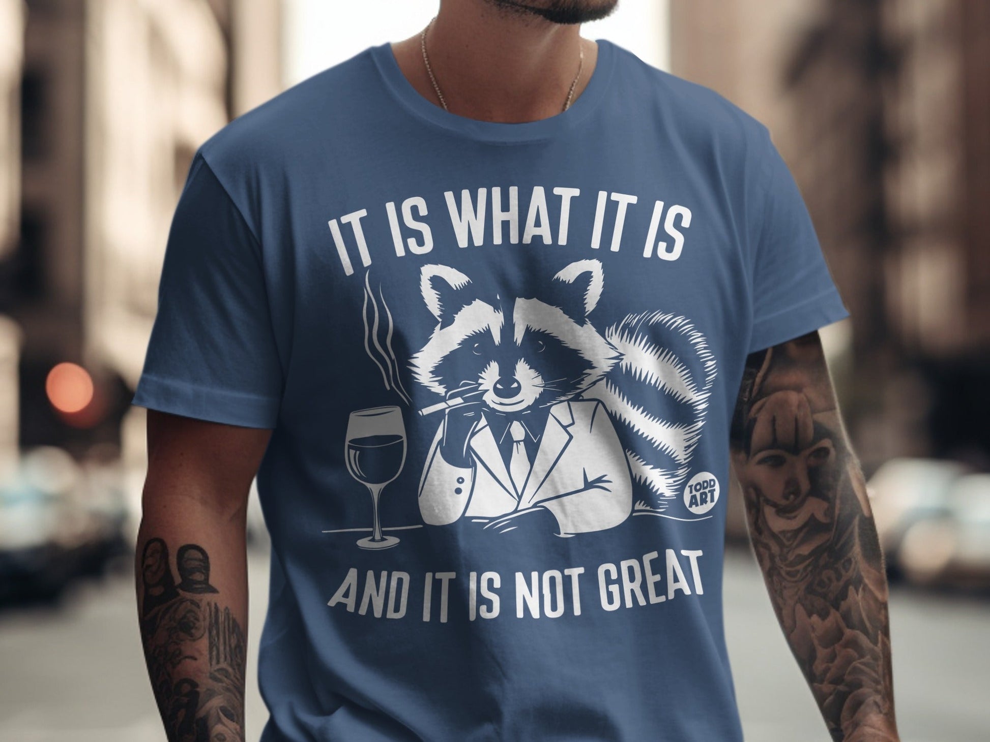 Blue t-shirt with a raccoon wearing a suit and smoking, text reads "IT IS WHAT IT IS AND IT IS NOT GREAT".