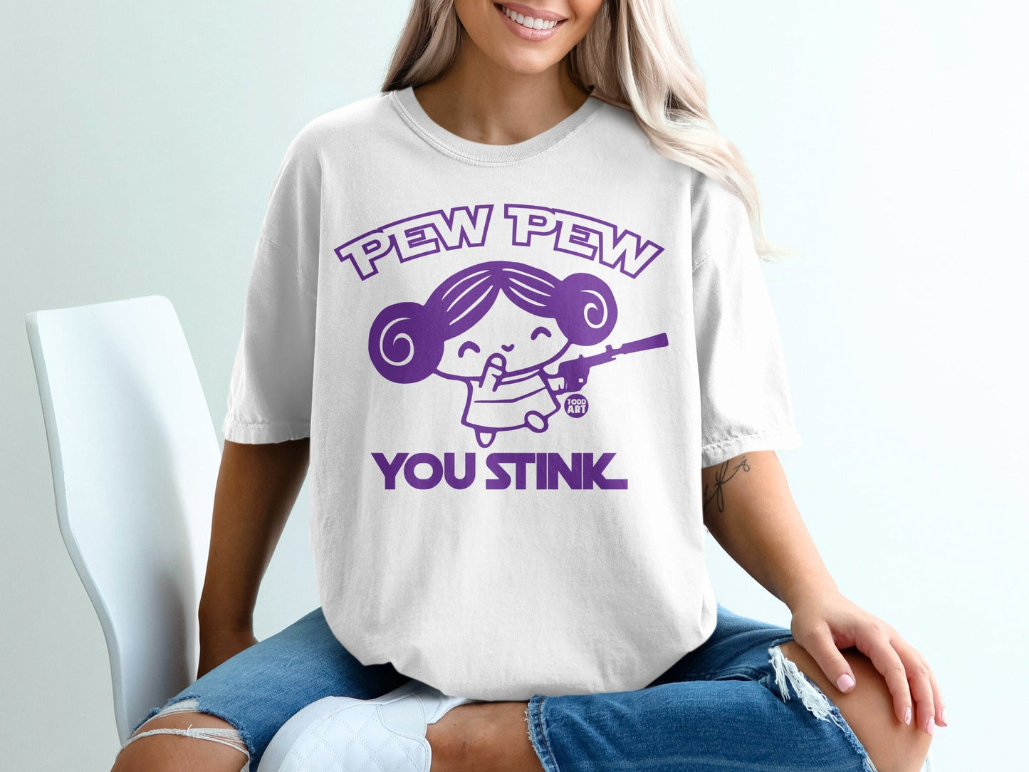 White t-shirt with purple graphic of a cartoon girl holding a gun, text "PEW PEW YOU STINK".