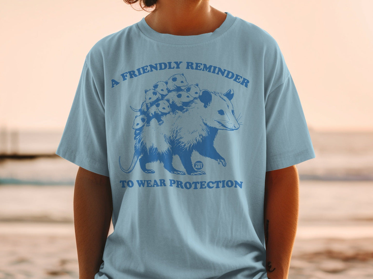 Light blue t-shirt with graphic of an opossum and babies, text "A friendly reminder to wear protection".