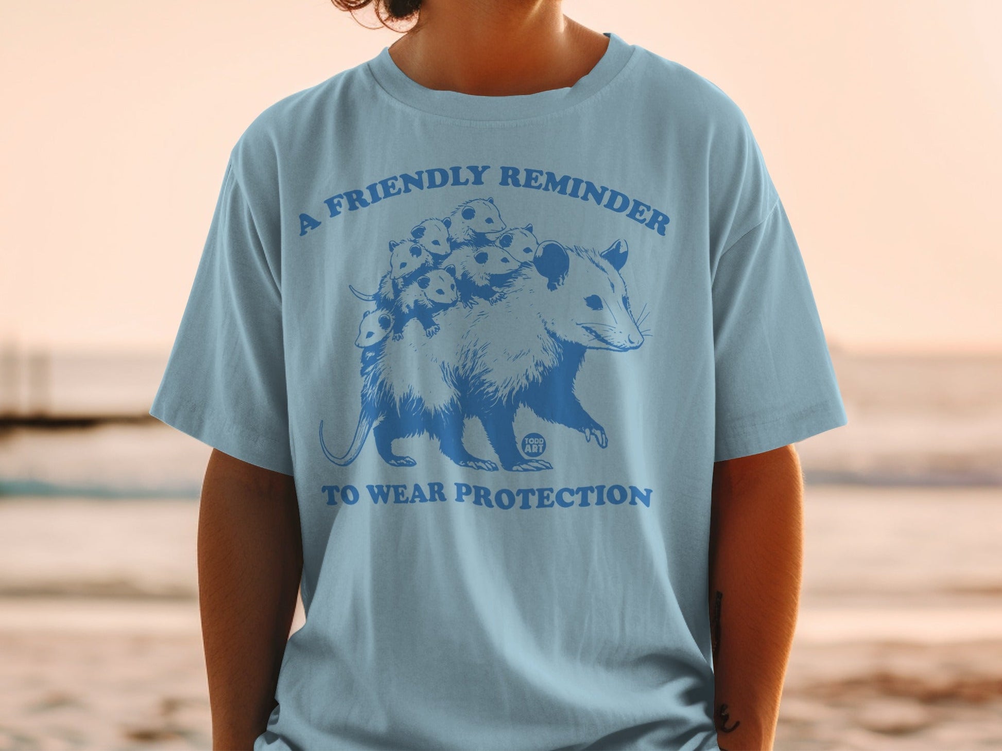 Light blue t-shirt with graphic of an opossum and babies, text "A friendly reminder to wear protection".
