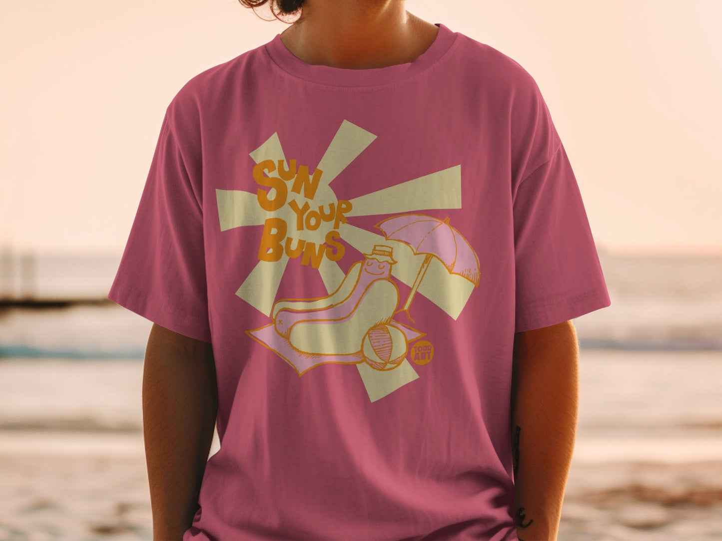 Pink t-shirt with cartoon hot dog wearing sunglasses and hat at beach.