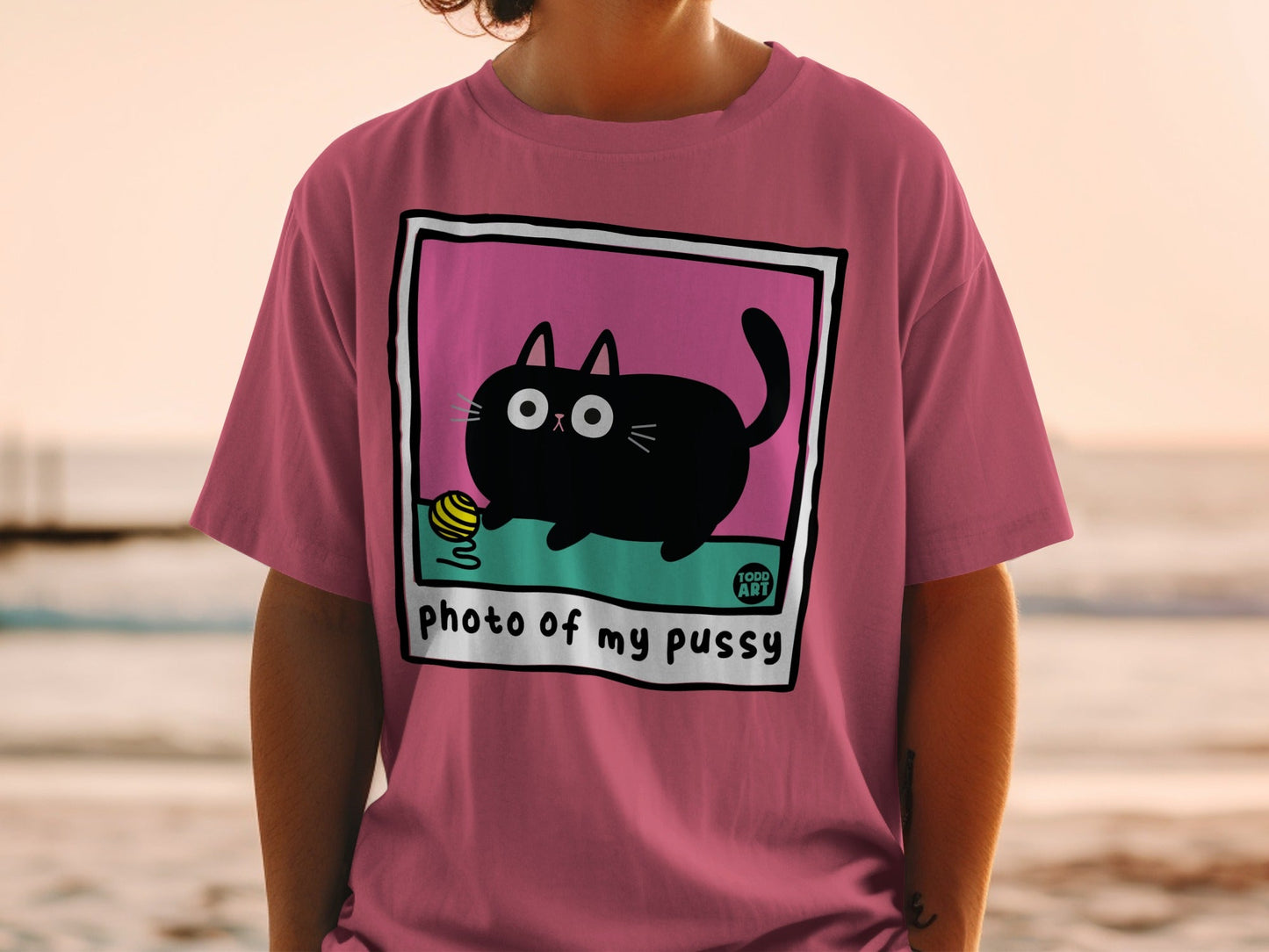 Pink t-shirt with cartoon cat playing with yellow yarn, text "photo of my pussy".
