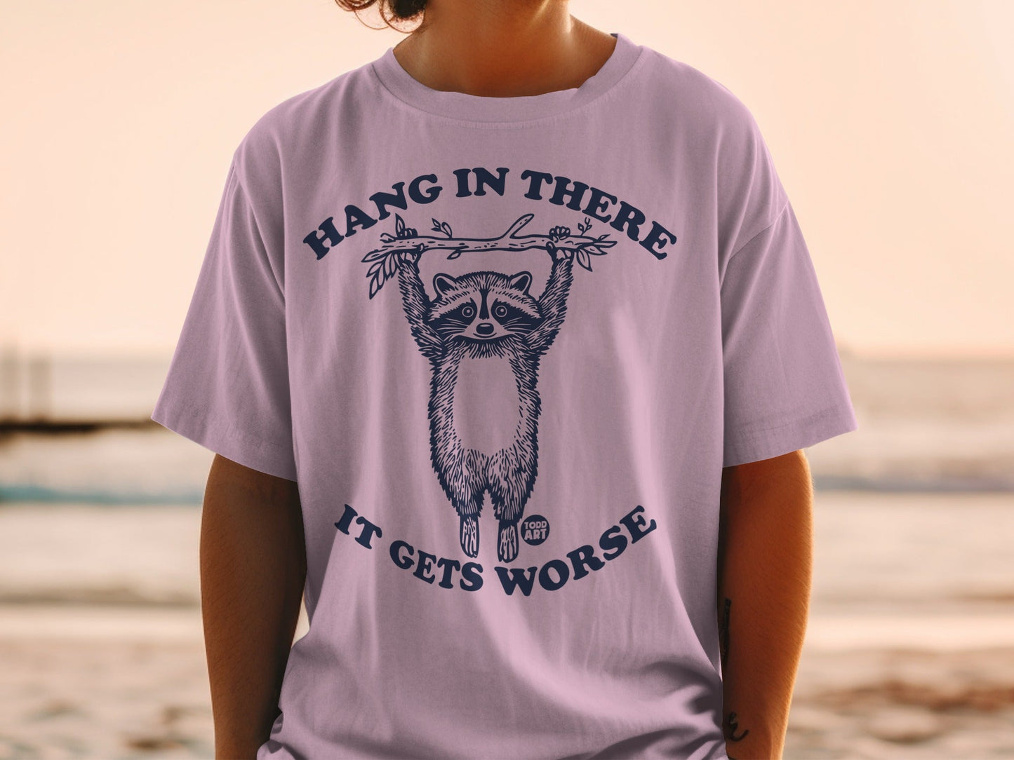 T-shirt with raccoon hanging from branch, text "HANG IN THERE IT GETS WORSE".