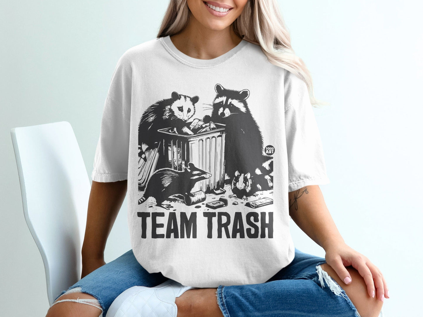 White t-shirt with a graphic of raccoons and an opossum rummaging through trash.