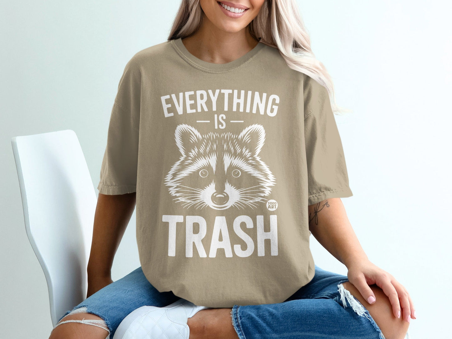 Woman wearing tan t-shirt with raccoon graphic and "EVERYTHING IS TRASH" text.