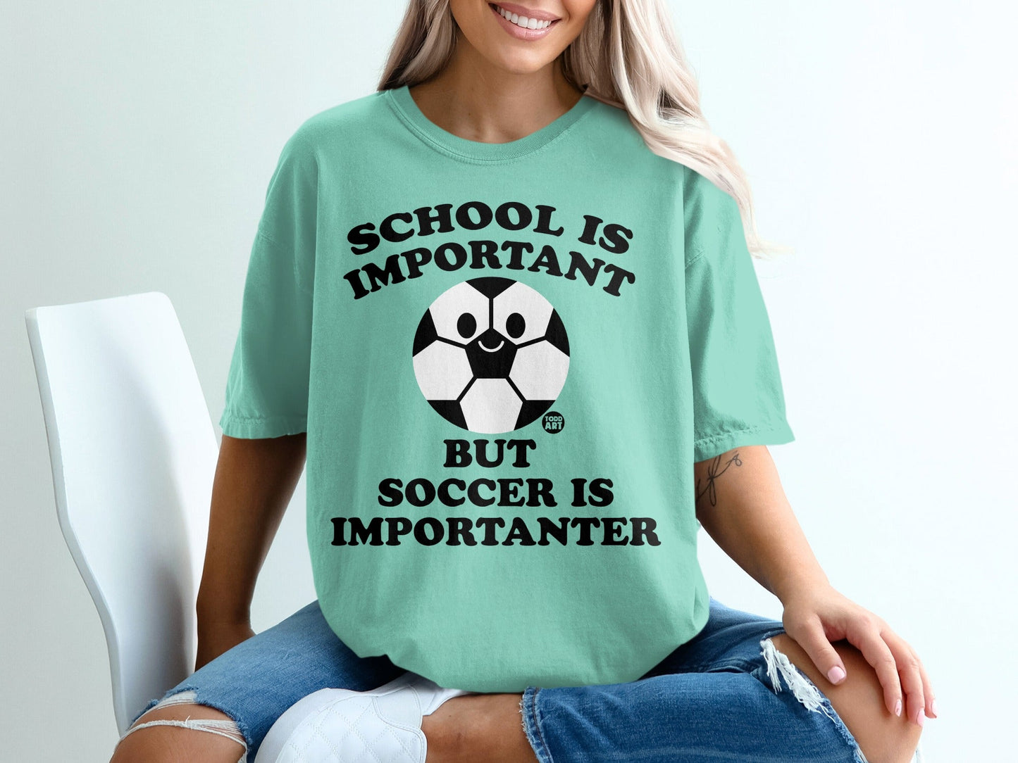 "School is important but soccer is importanter" text on a mint green t-shirt with a smiling soccer ball.