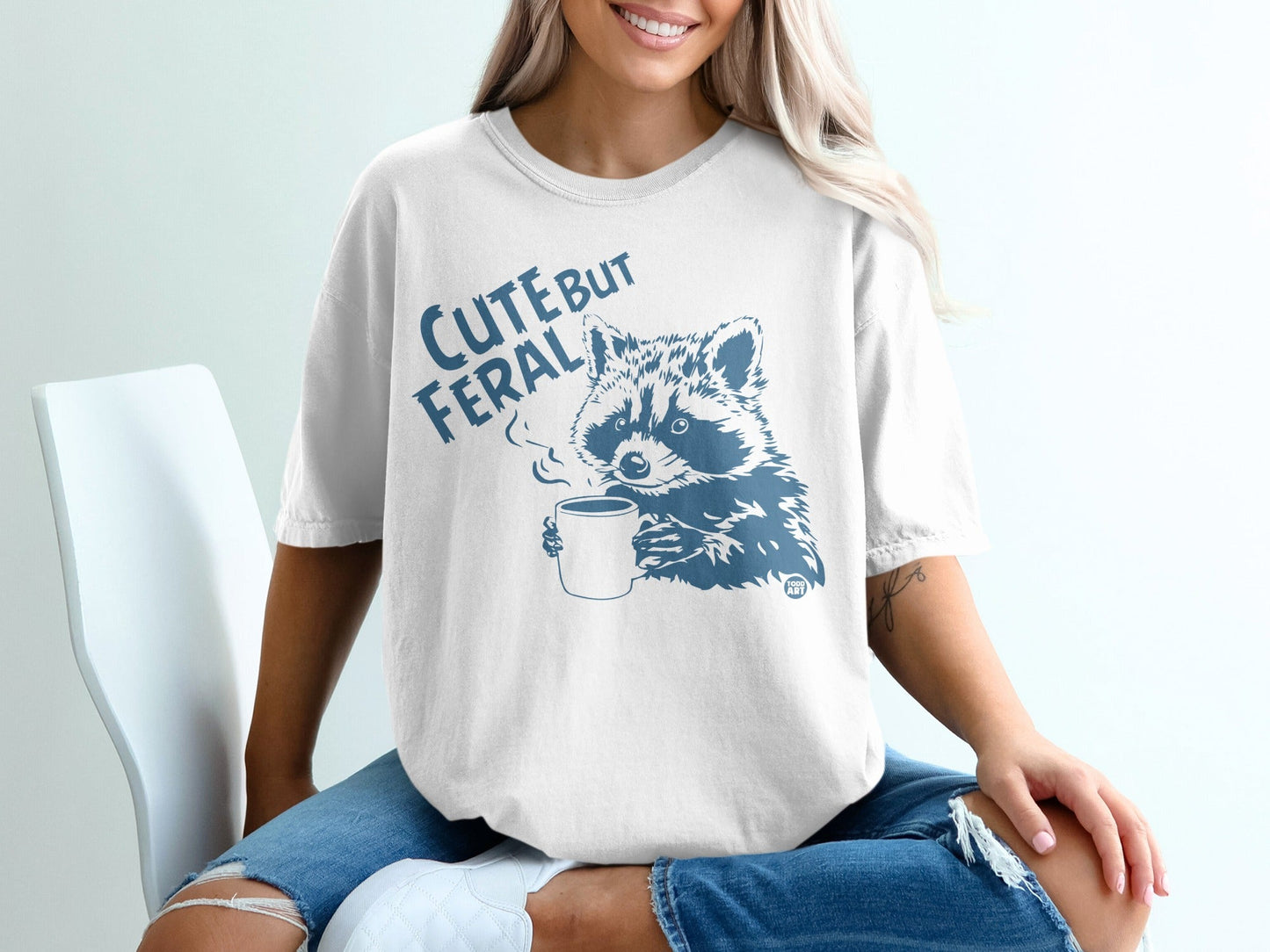 White t-shirt with "Cute But Feral" and a raccoon drinking coffee graphic.