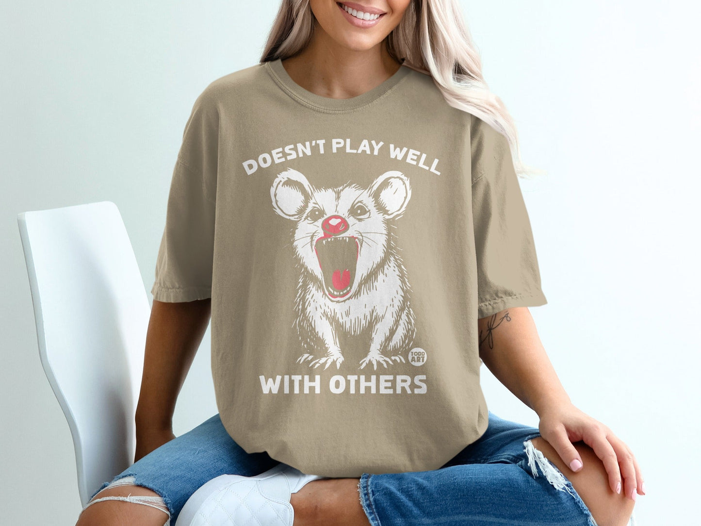 Tan t-shirt with graphic of opossum and text "DOESN'T PLAY WELL WITH OTHERS".