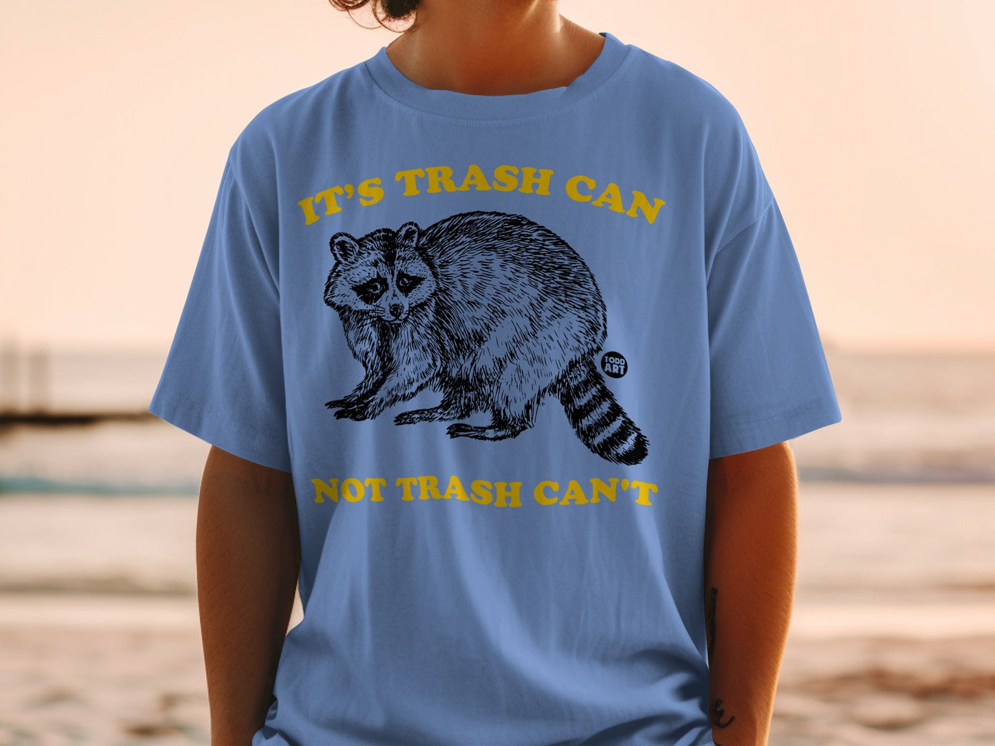 Blue t-shirt with a raccoon graphic and text "IT'S TRASH CAN NOT TRASH CAN'T".