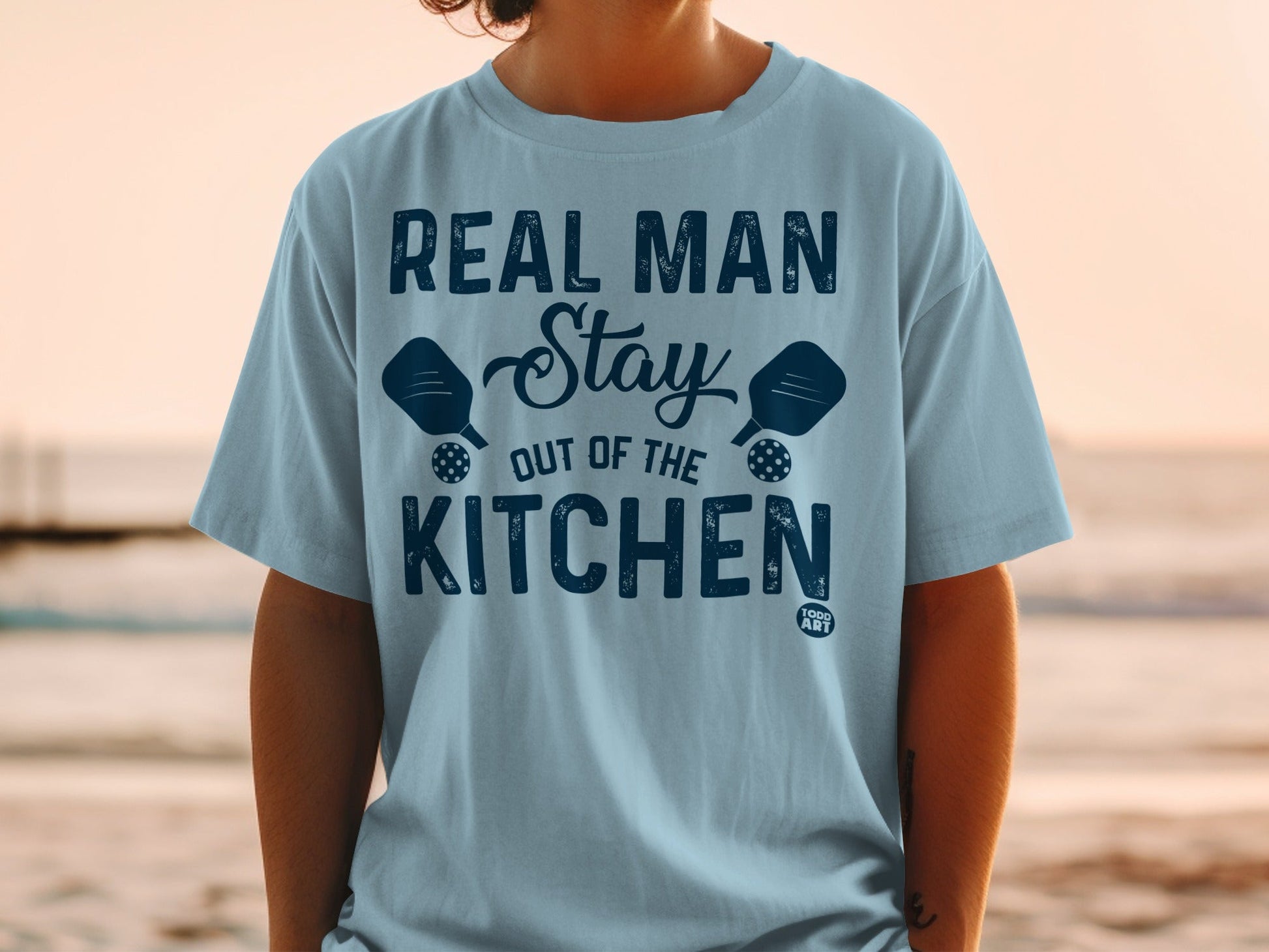 Light blue t-shirt with "Real Man Stay Out of the Kitchen" text and pickleball paddles.