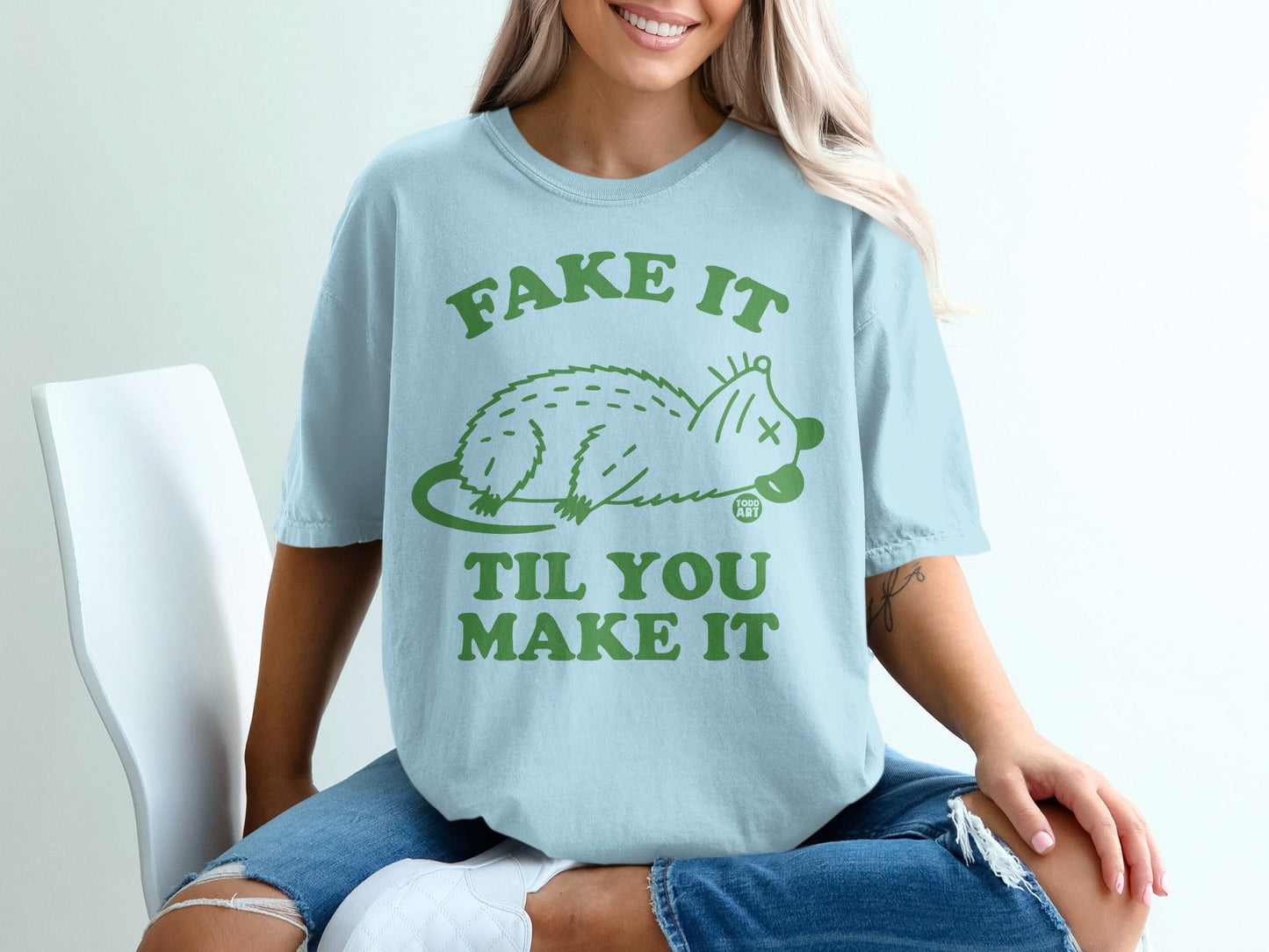 Light blue t-shirt with green text "Fake It Til You Make It" and a cartoon opossum.