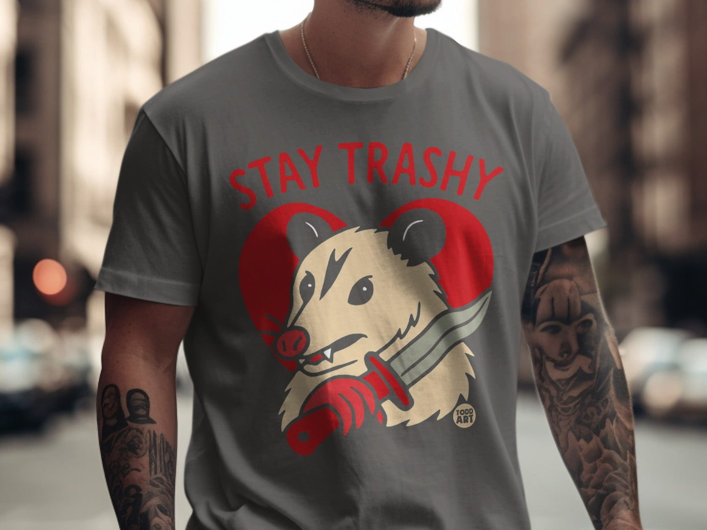 Gray t-shirt with a possum holding a knife inside a red heart, reads "STAY TRASHY".