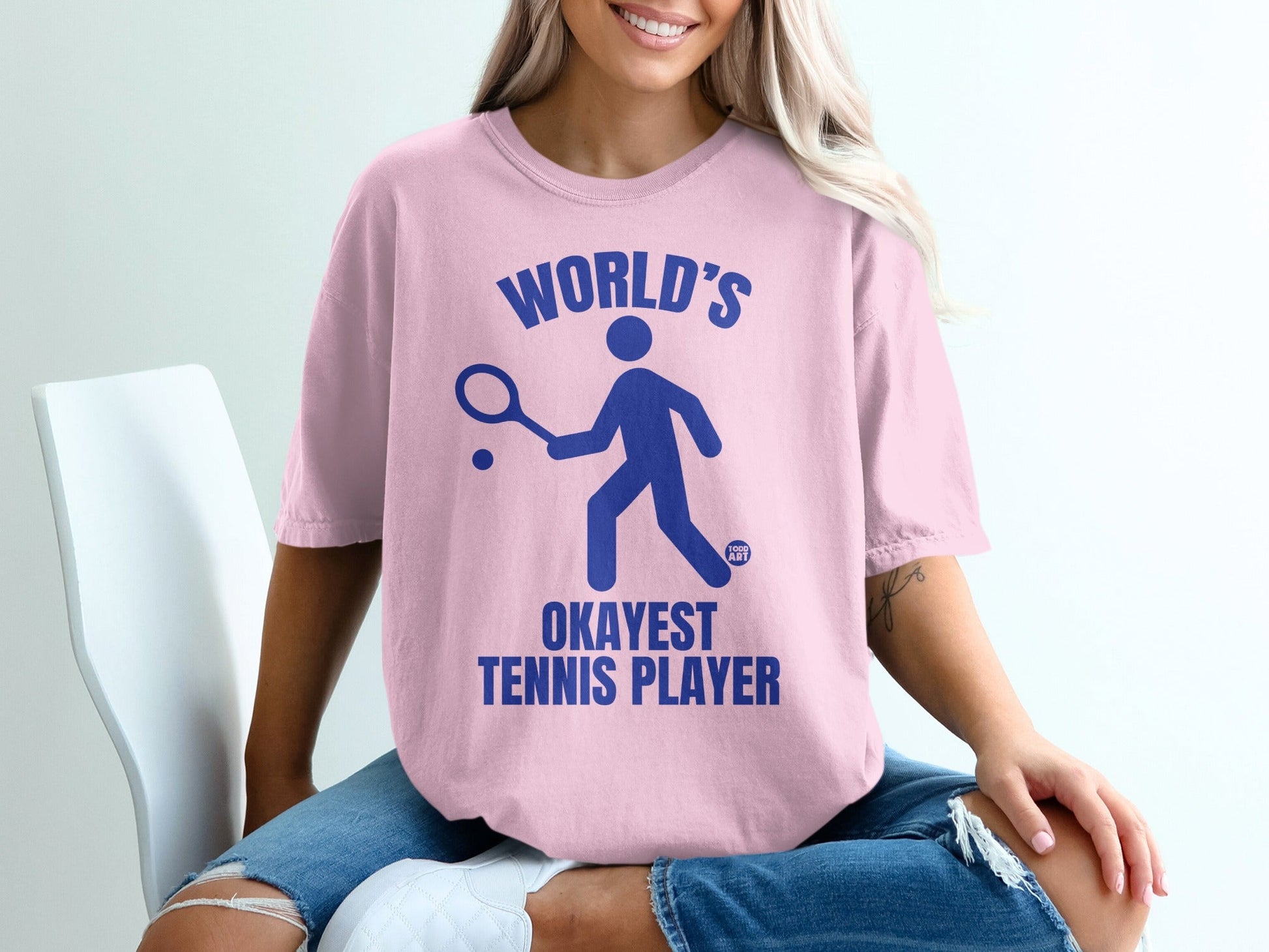 Pink t-shirt with blue text and graphic: "World's Okayest Tennis Player."