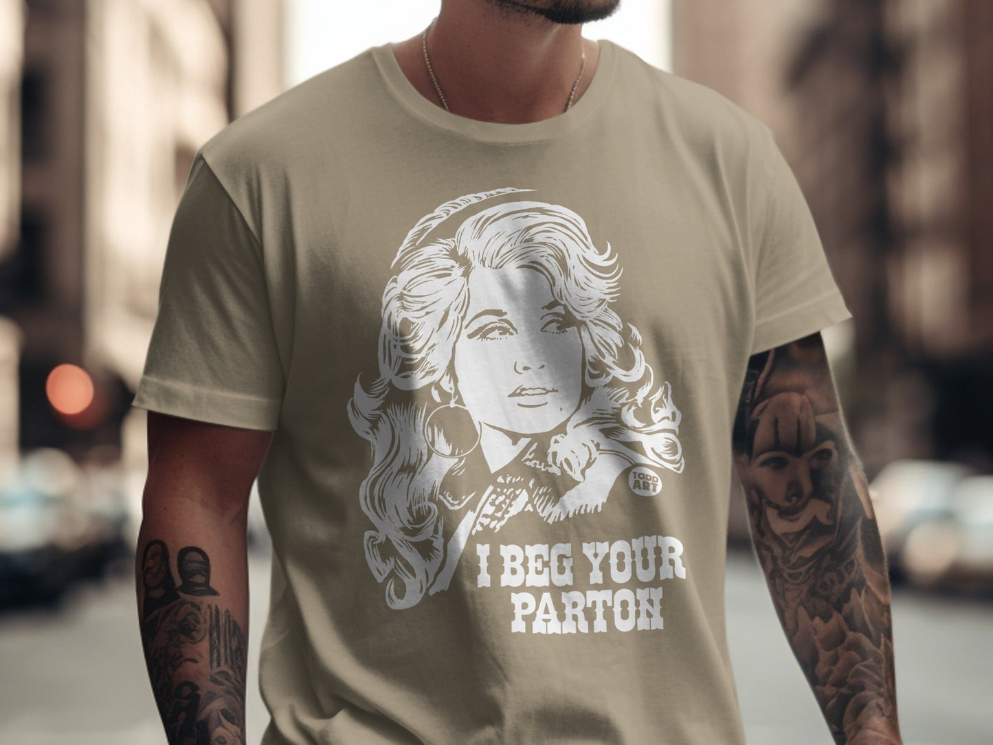 Tan t-shirt with a graphic of Dolly Parton and text "I BEG YOUR PARTON".