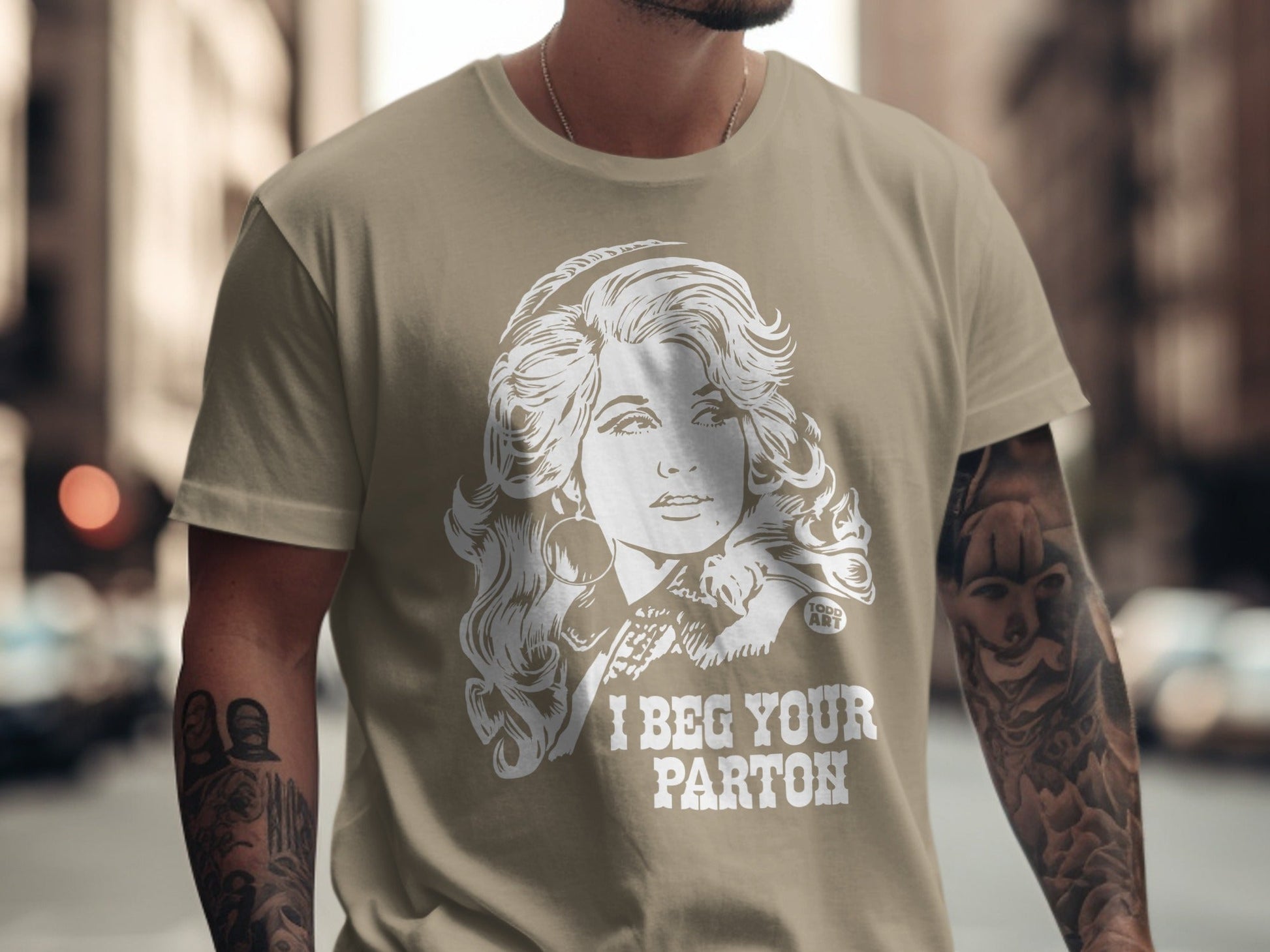 Tan t-shirt with a graphic of Dolly Parton and text "I BEG YOUR PARTON".