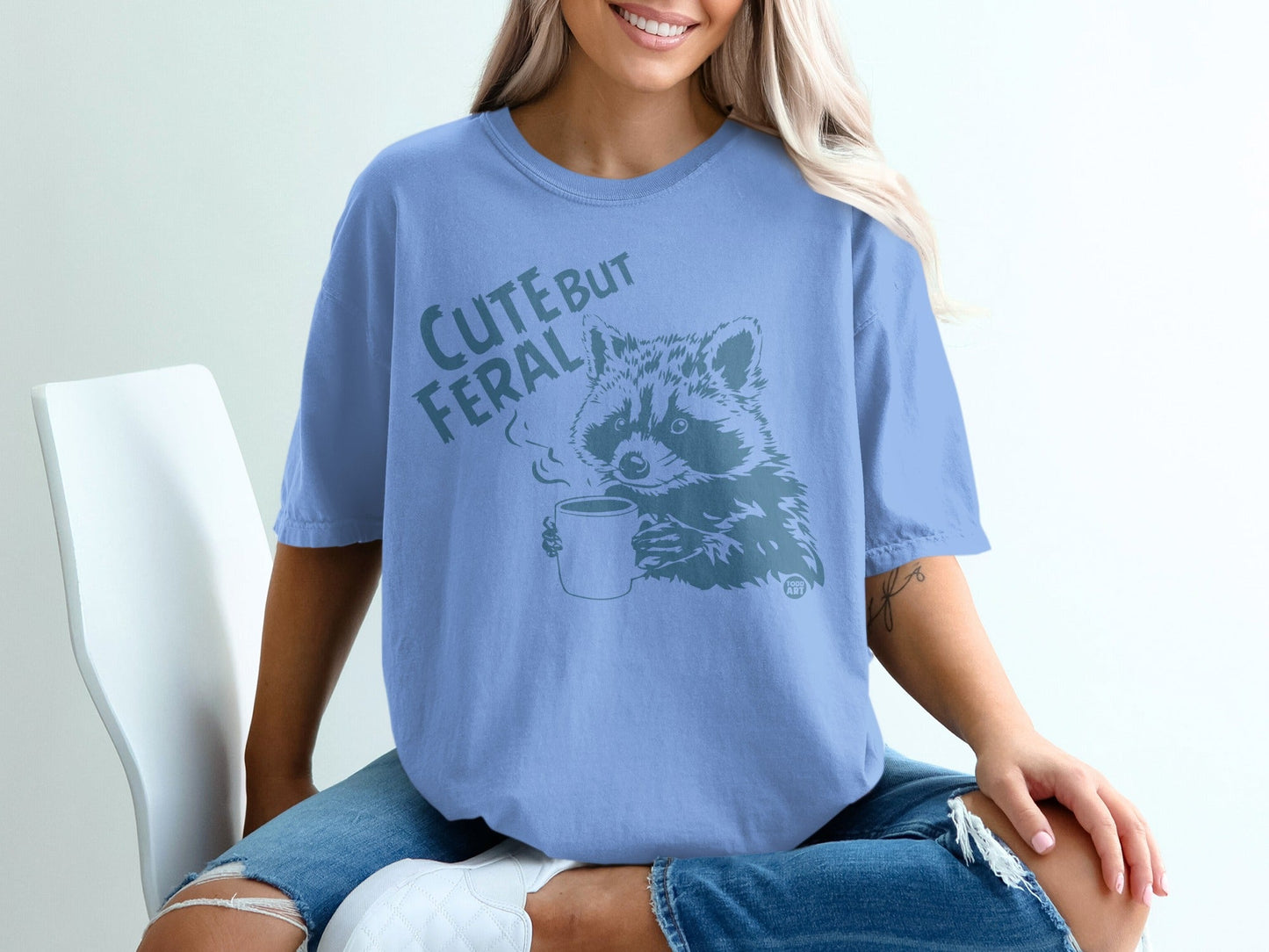 Blue t-shirt with a raccoon holding a mug and text that says "Cute but Feral".