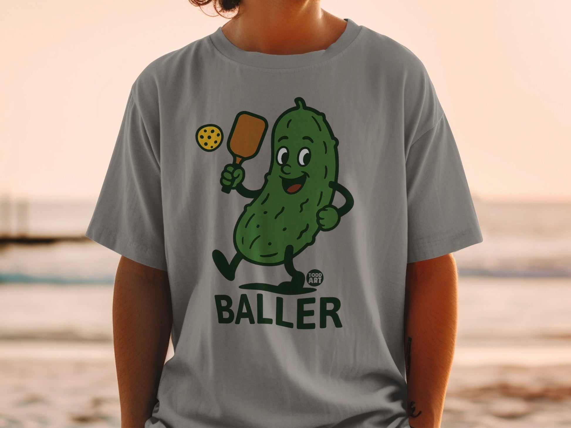 Gray t-shirt with cartoon pickle playing pickleball, "GOOD ART" logo.