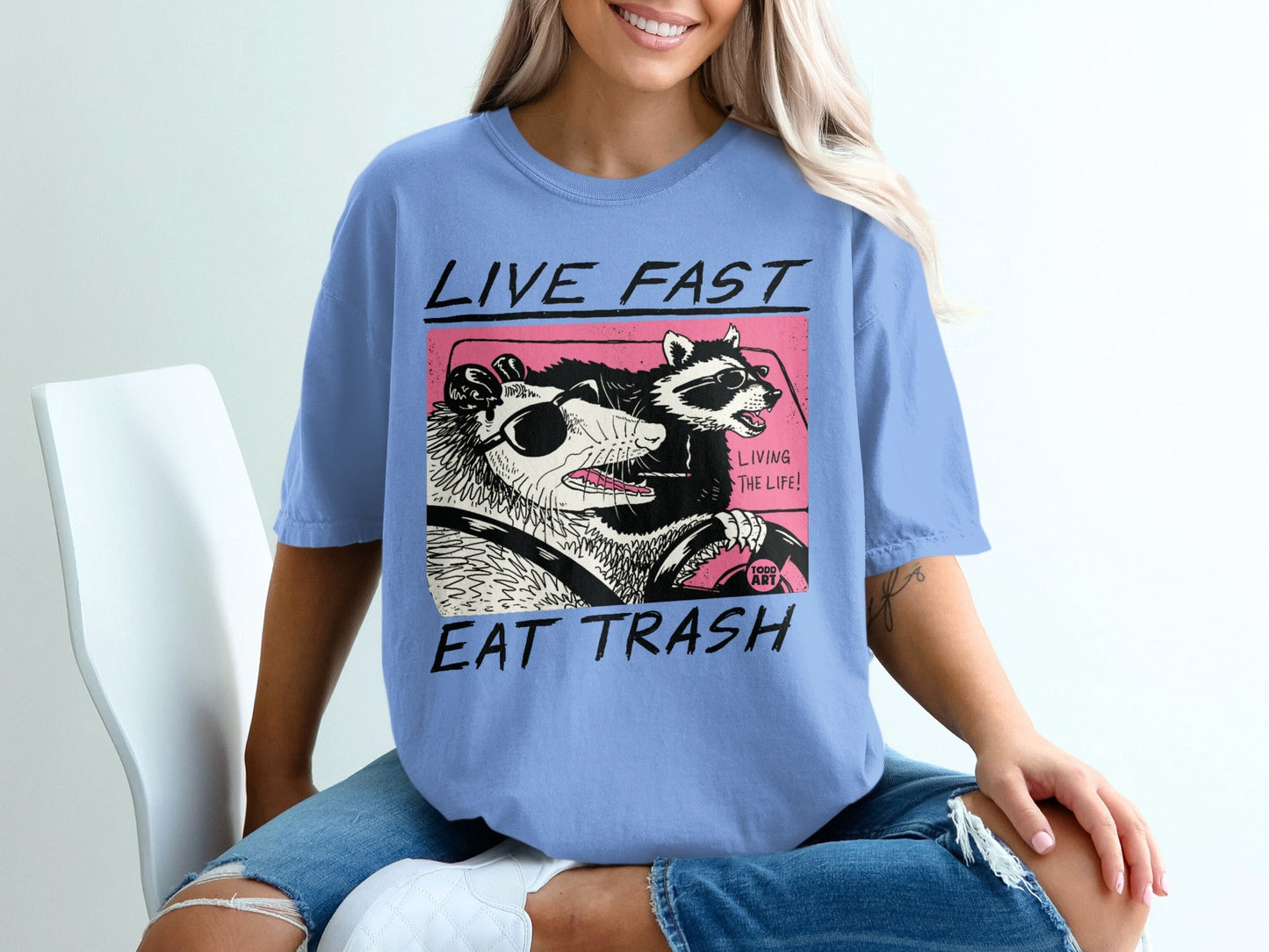 T-shirt graphic of a raccoon and possum driving, with text "LIVE FAST EAT TRASH LIVING THE LIFE!".