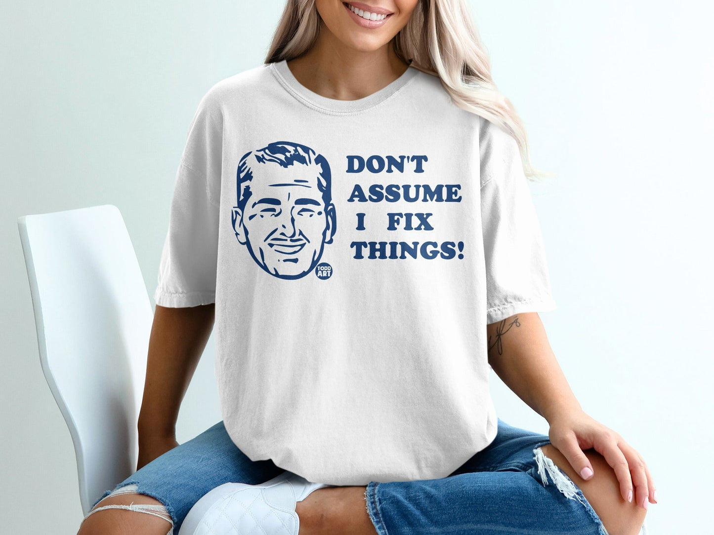 White t-shirt with blue text "DON'T ASSUME I FIX THINGS!" and cartoon man.
