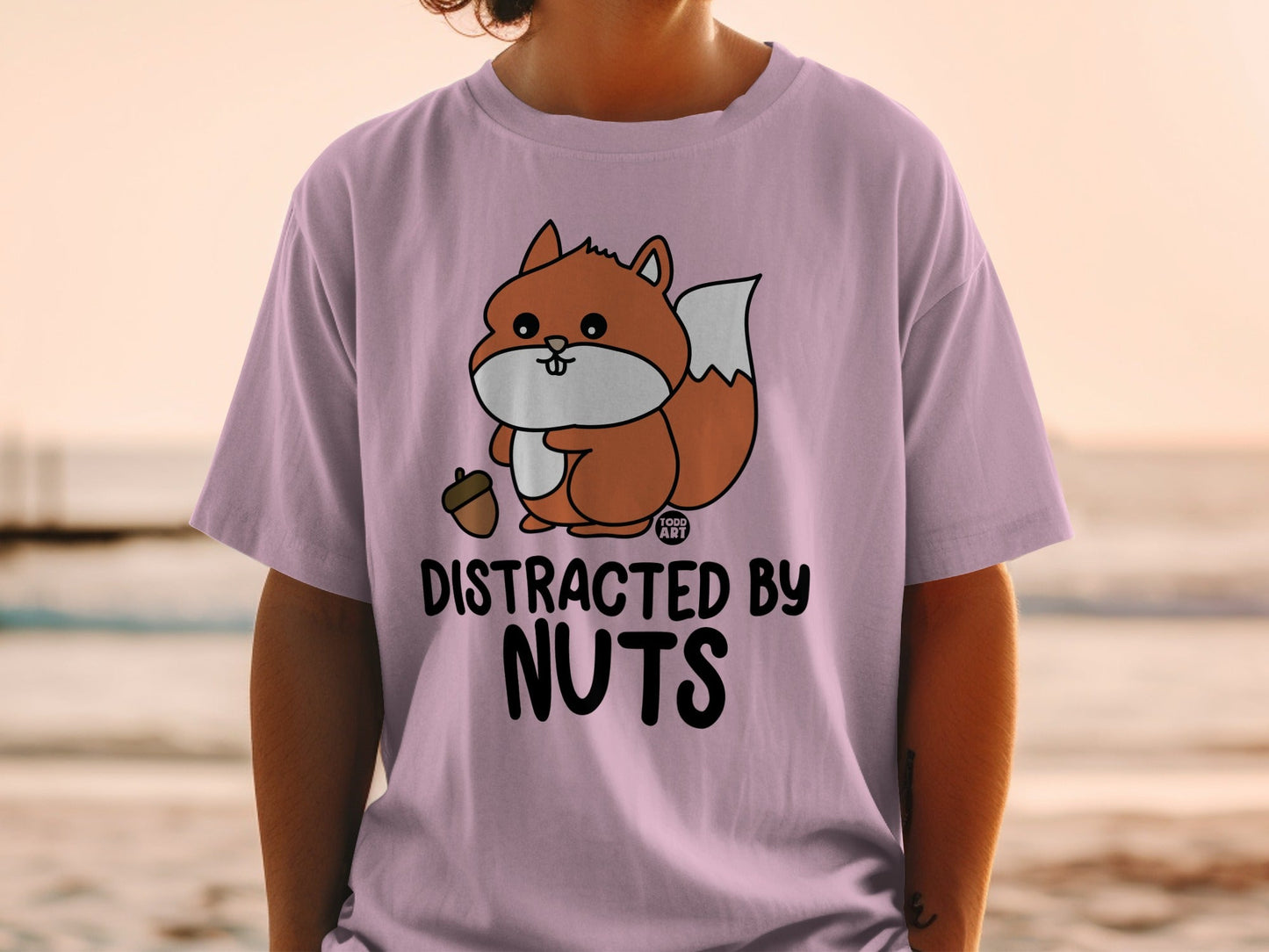 Cartoon squirrel holding an acorn on a pink t-shirt with "DISTRACTED BY NUTS" text.