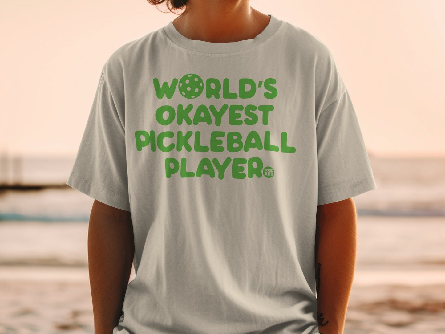 Light grey t-shirt with green text "WORLD'S OKAYEST PICKLEBALL PLAYER".