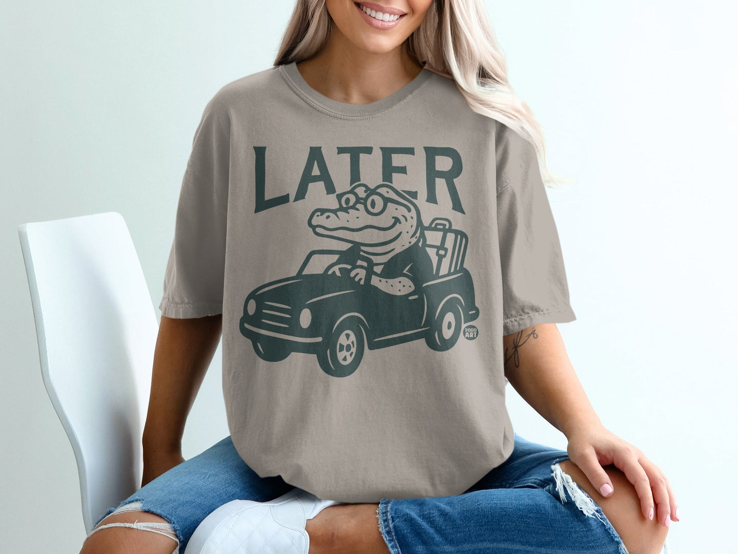 Green graphic of alligator driving a car on a taupe t-shirt.