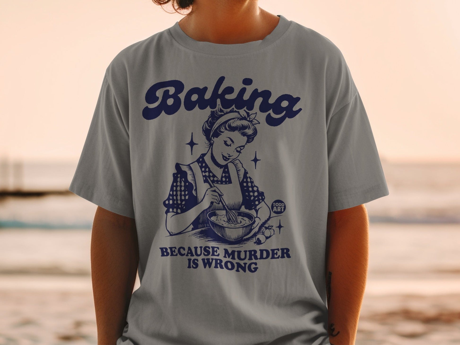 Grey t-shirt with retro graphic of woman baking and text "Baking because murder is wrong".