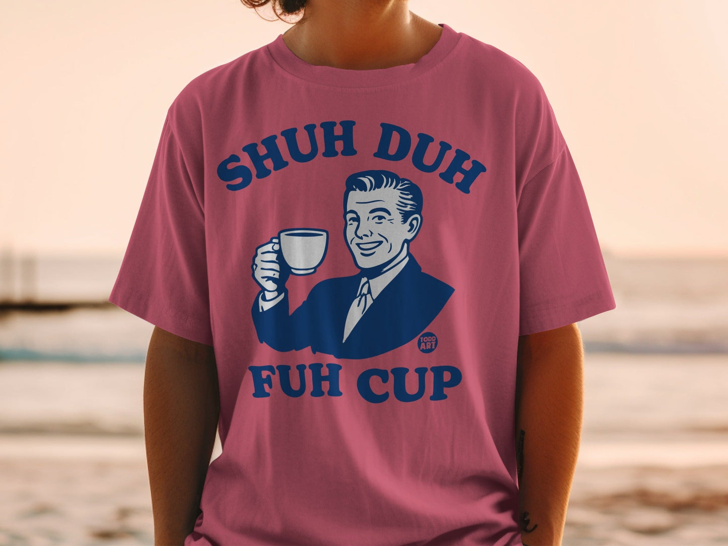 Pink t-shirt with blue text "SHUH DUH FUH CUP" and man holding coffee.